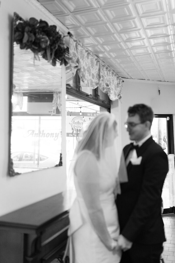 black and white image holding hands and smiling at each other, out of focus in front of an interior mirror (in focus), reflecting the word "Anthony's" from an exterior window