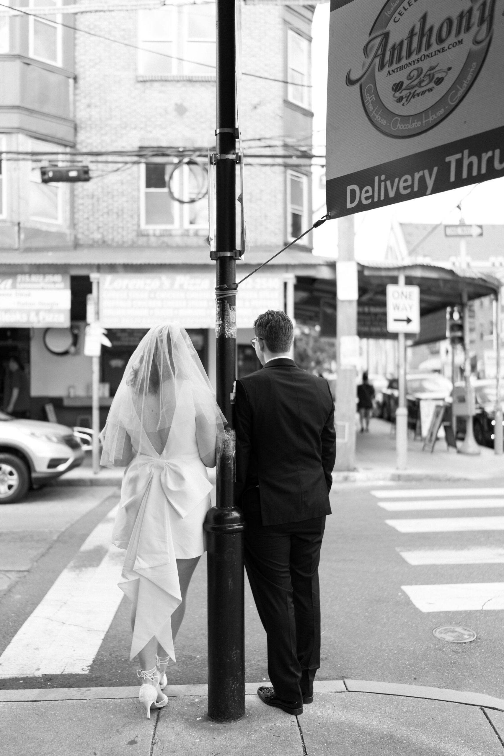 black and white image of a bride and a groom leaning on opposite sides of a lamp post, facing toward the road away from camera, waiting for an Uber to pick them up