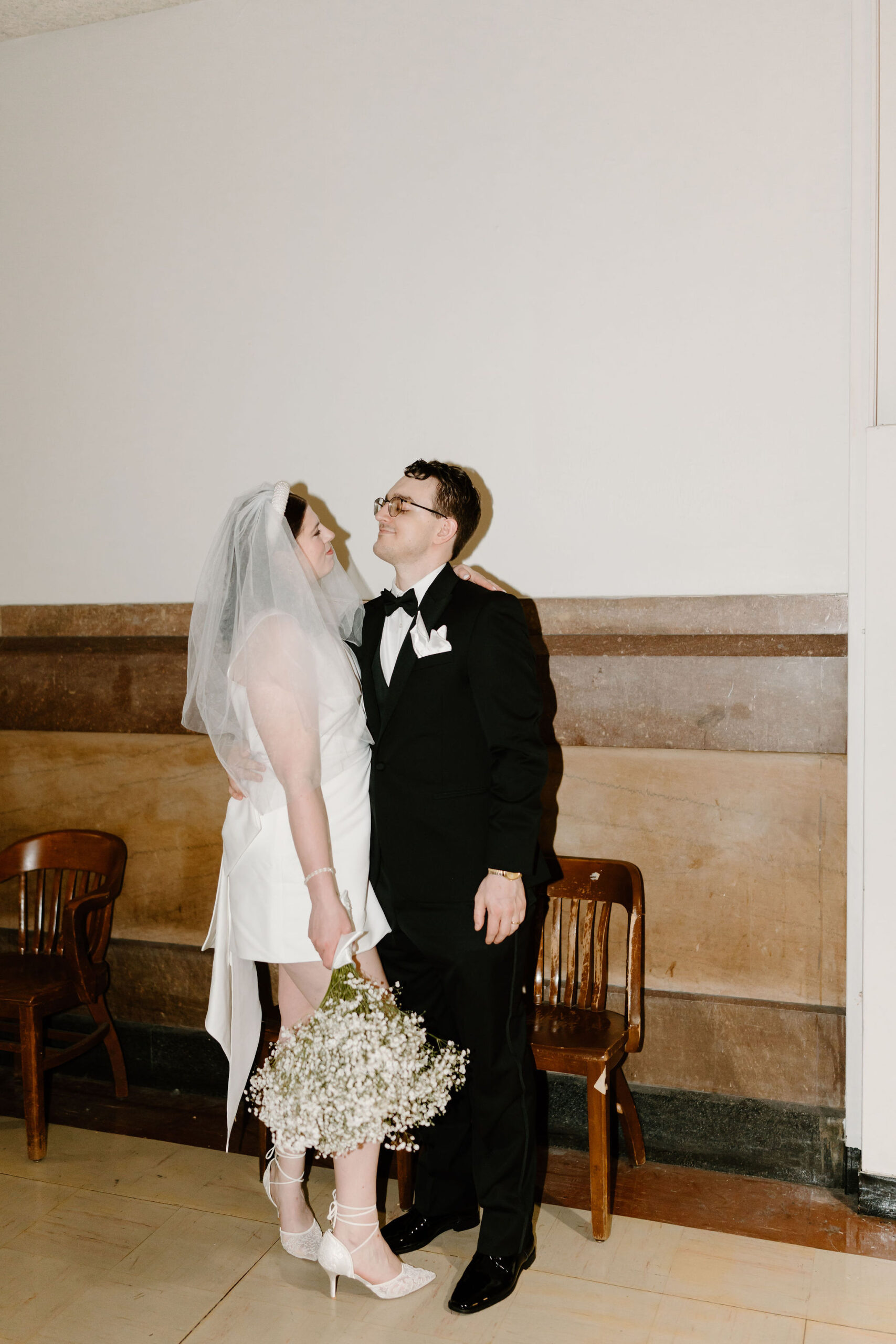 bride and groom embracing and smiling at each other inside city hall hallway, bride holding her bouquet down at her side