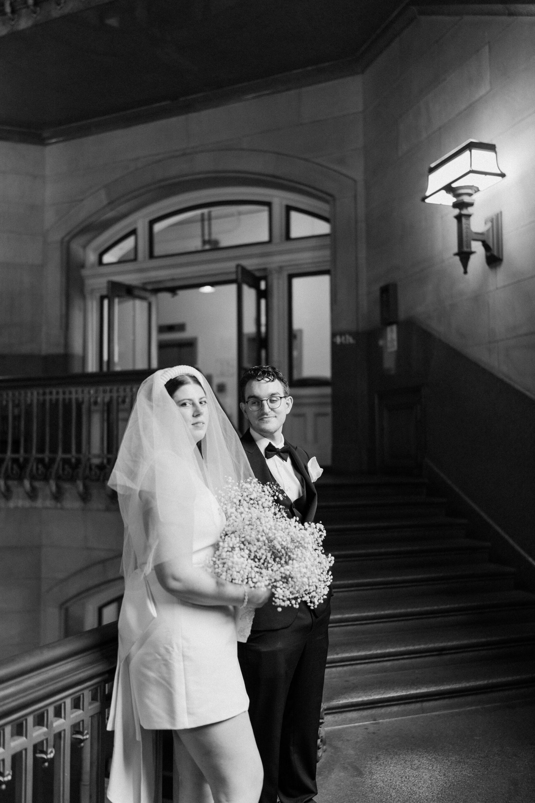 black and white image of bride and groom leaning against a railing in a winding stairwell, looking toward the camera