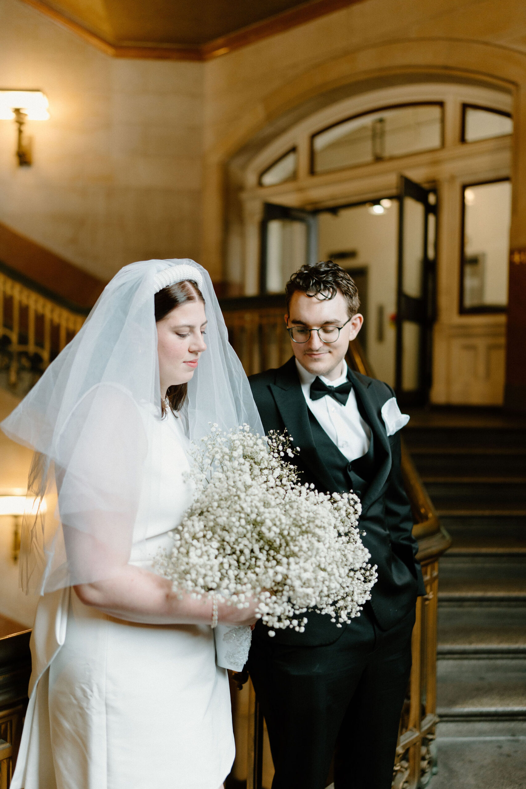 bride and groom leaning on railing in a large stairwell, both looking down at bride's bouquet of baby's breath