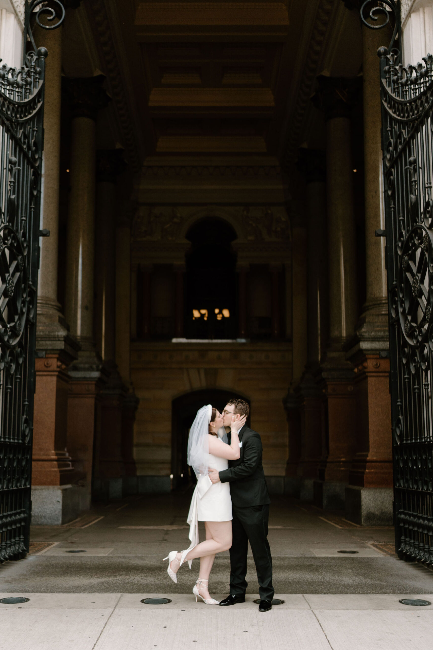 groom holding bride around the waist and kissing her, while she holds his face and pops up her right heel, as they stand in a large corridor with big metal gates