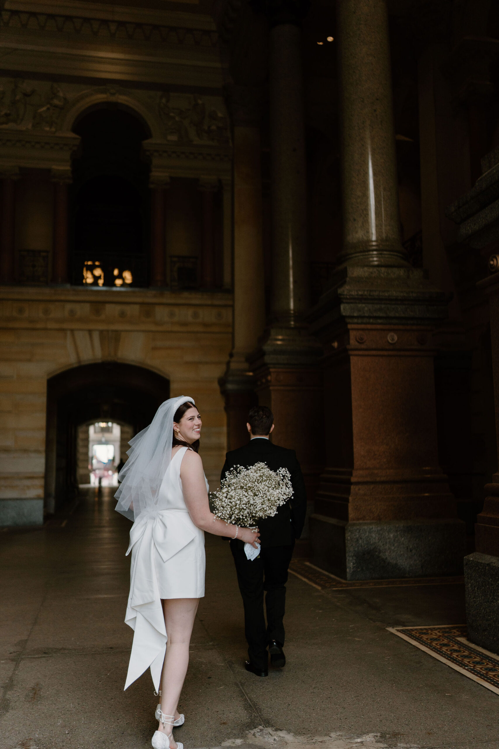 bride looking over her shoulder as she and groom walk through a large stone corridor with interesting architecture