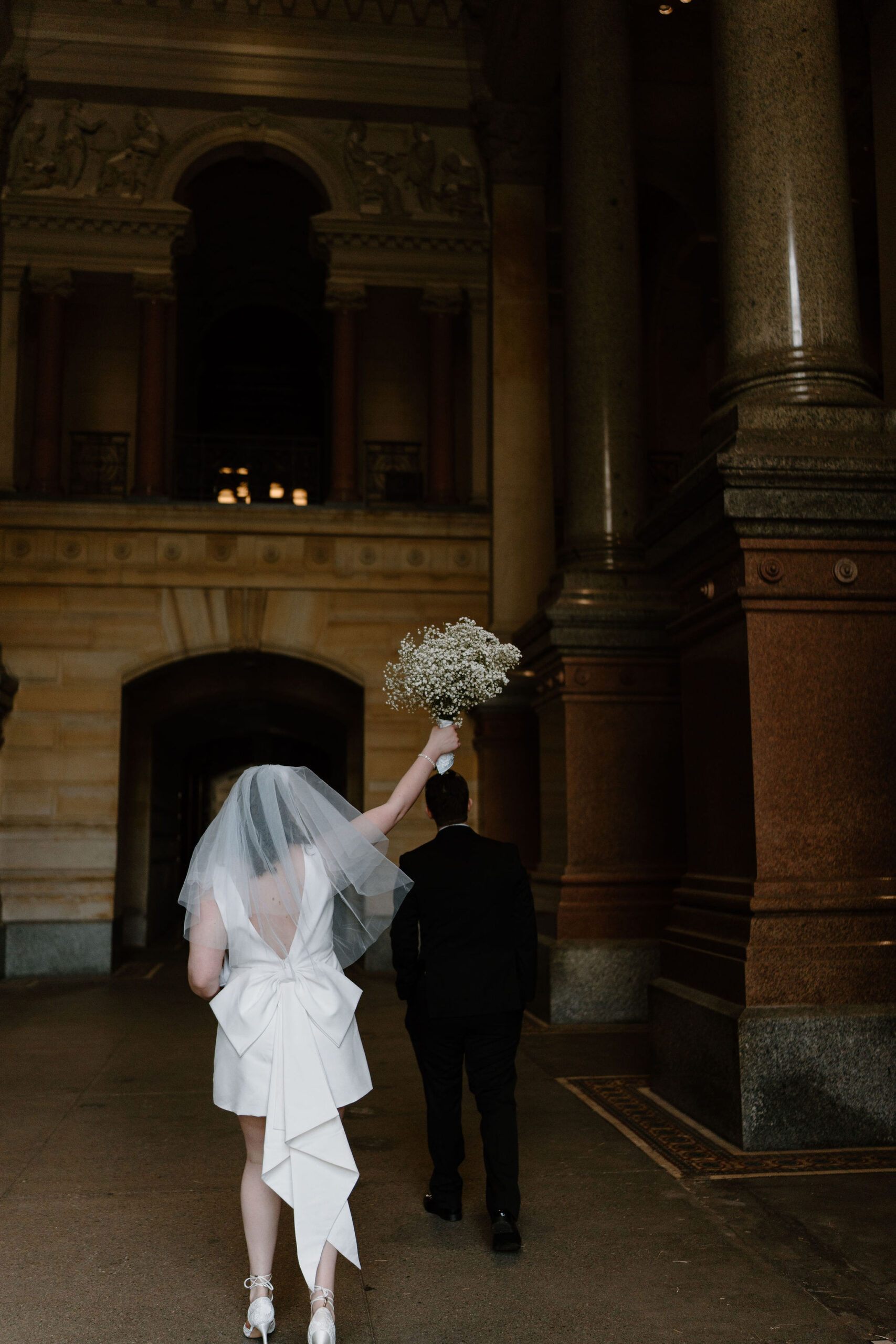 bride holding her flowers up over her head in a celebratory fashion as she and groom walk through a large stone corridor with interesting architecture