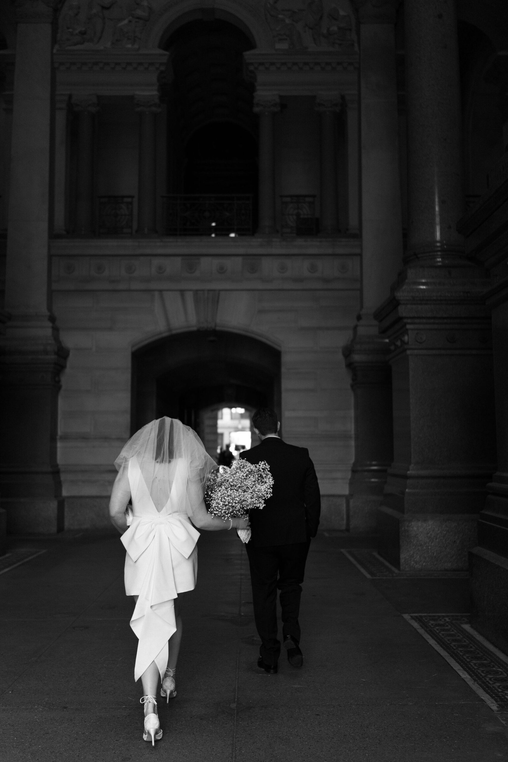 bride holding her flowers down at her side as she and groom walk through a large stone corridor with interesting architecture