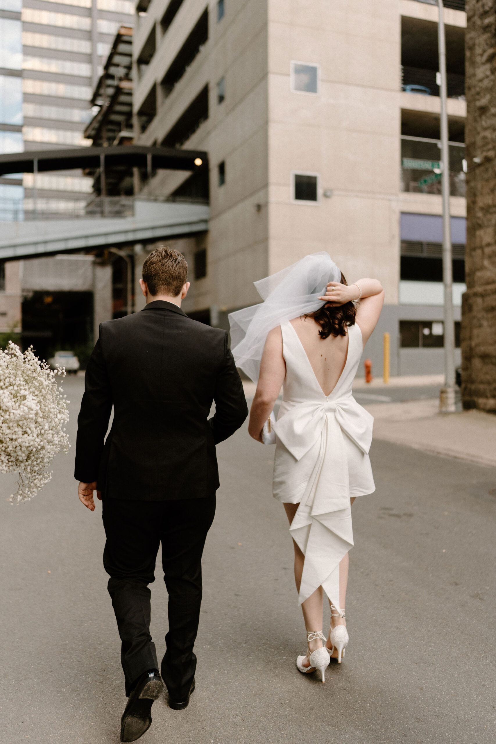 Bride and groom walking down the street toward their venue, bride holding her veil as it blows in the wind