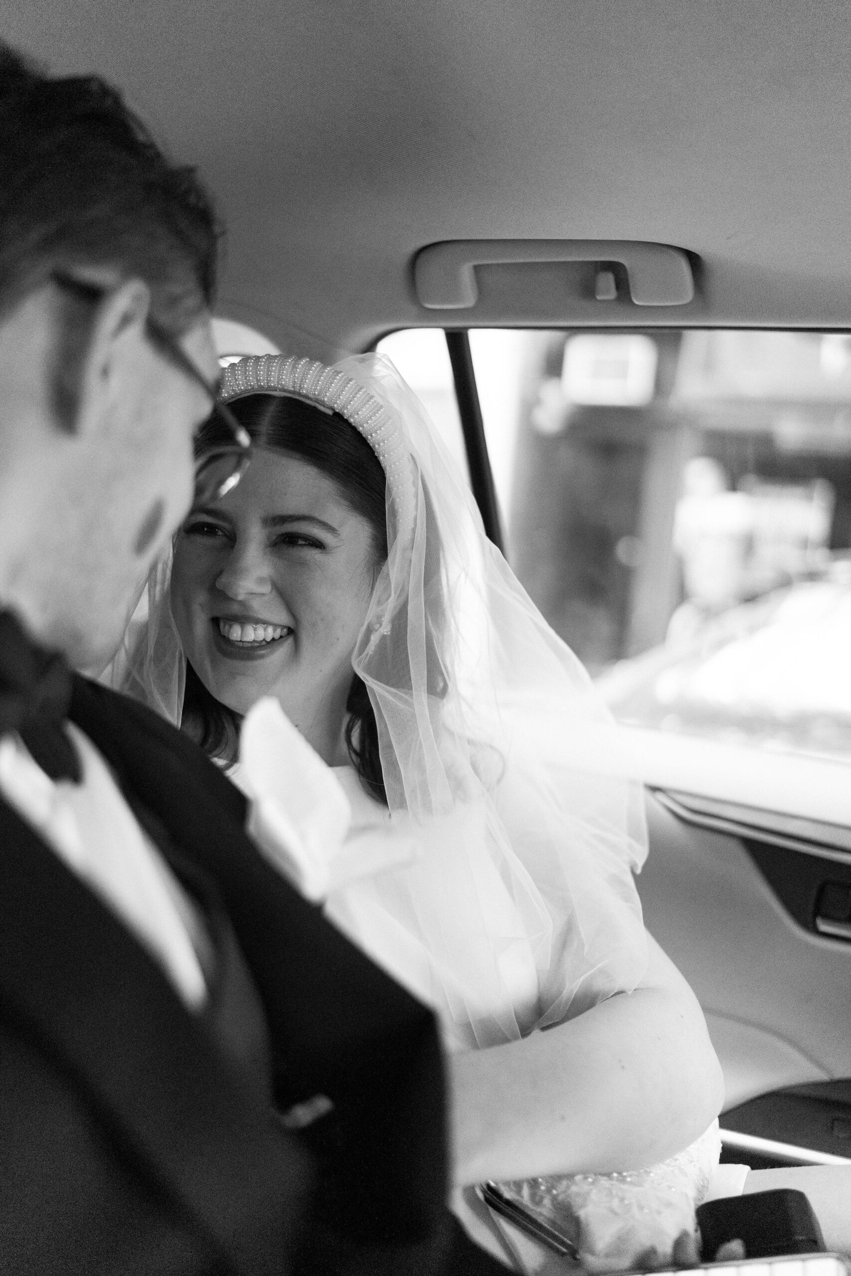 black and white image of bride and groom inside an Uber, smiling at each other and sitting closely together