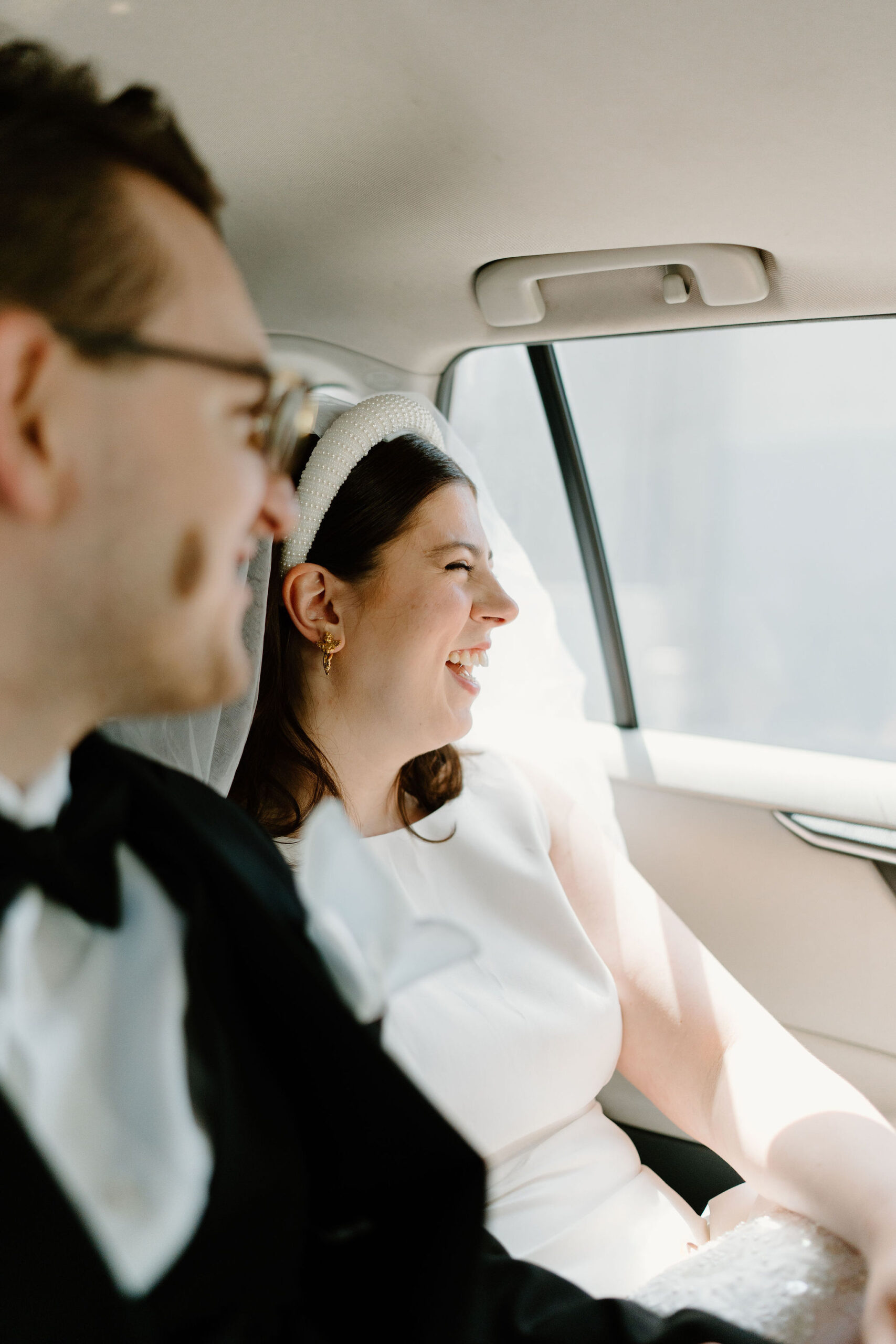 bride and groom seated next to each other in the back of a moving car, both laughing and smiling out the window