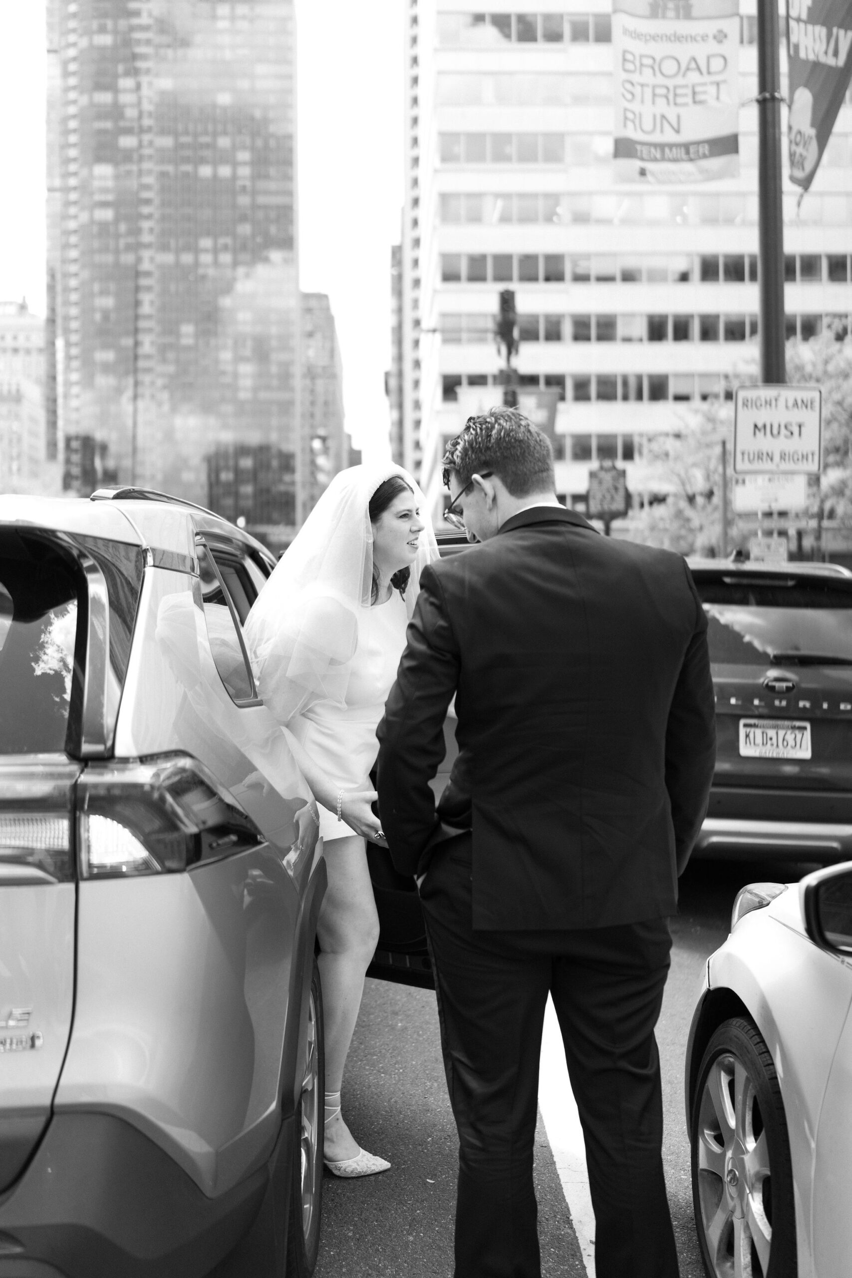 black and white image of bride (headband veil, short white dress) stepping out of a car as she arrives at her ceremony, groom next to her