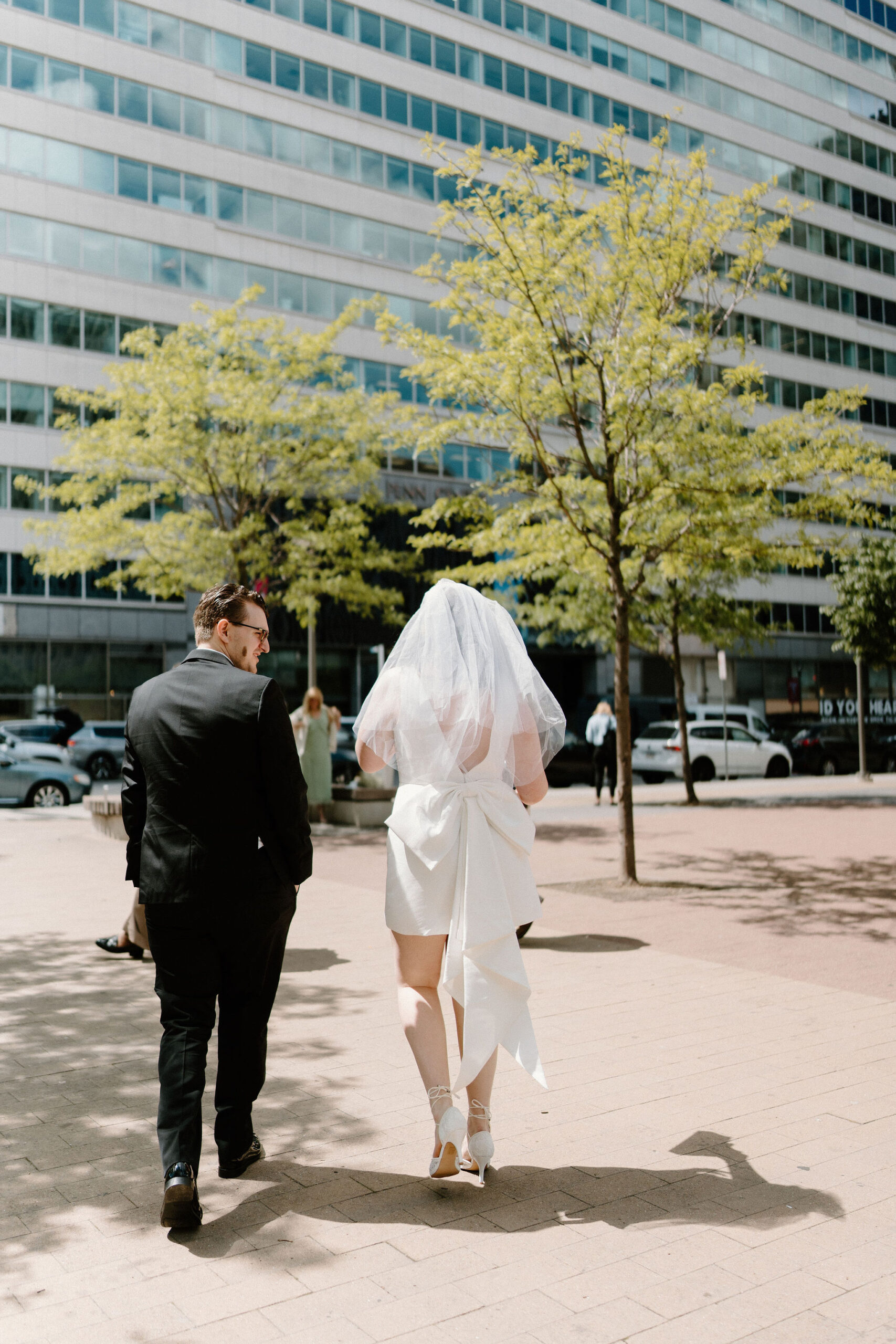 bride (short white dress with large bow in the back) and groom (black suit, glasses) walking up to their family, waiting for them in Love Park