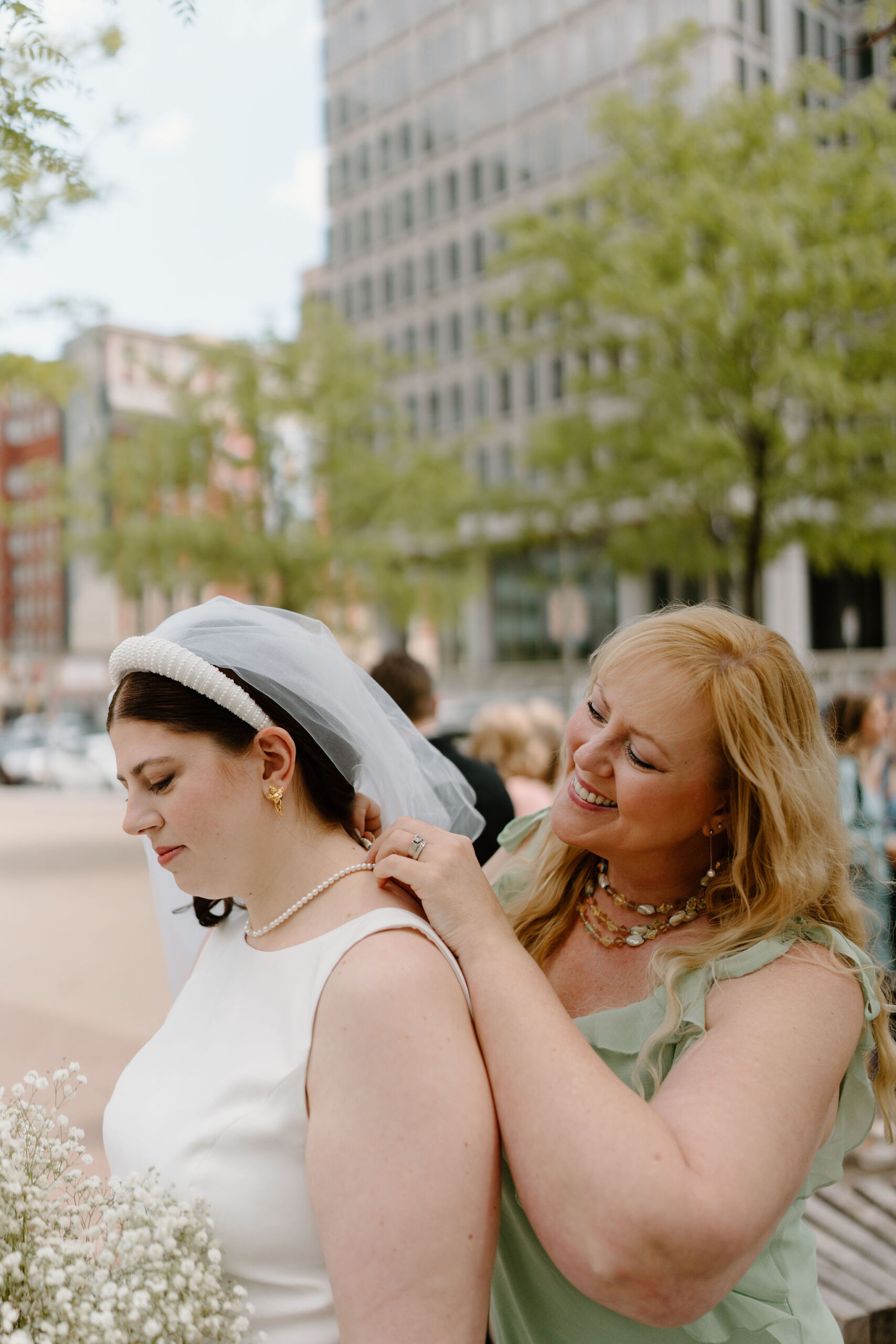 bride's mother (blonde hair, green dress) helping bride (white headband veil, holding baby's breath bouquet) to put on a necklace