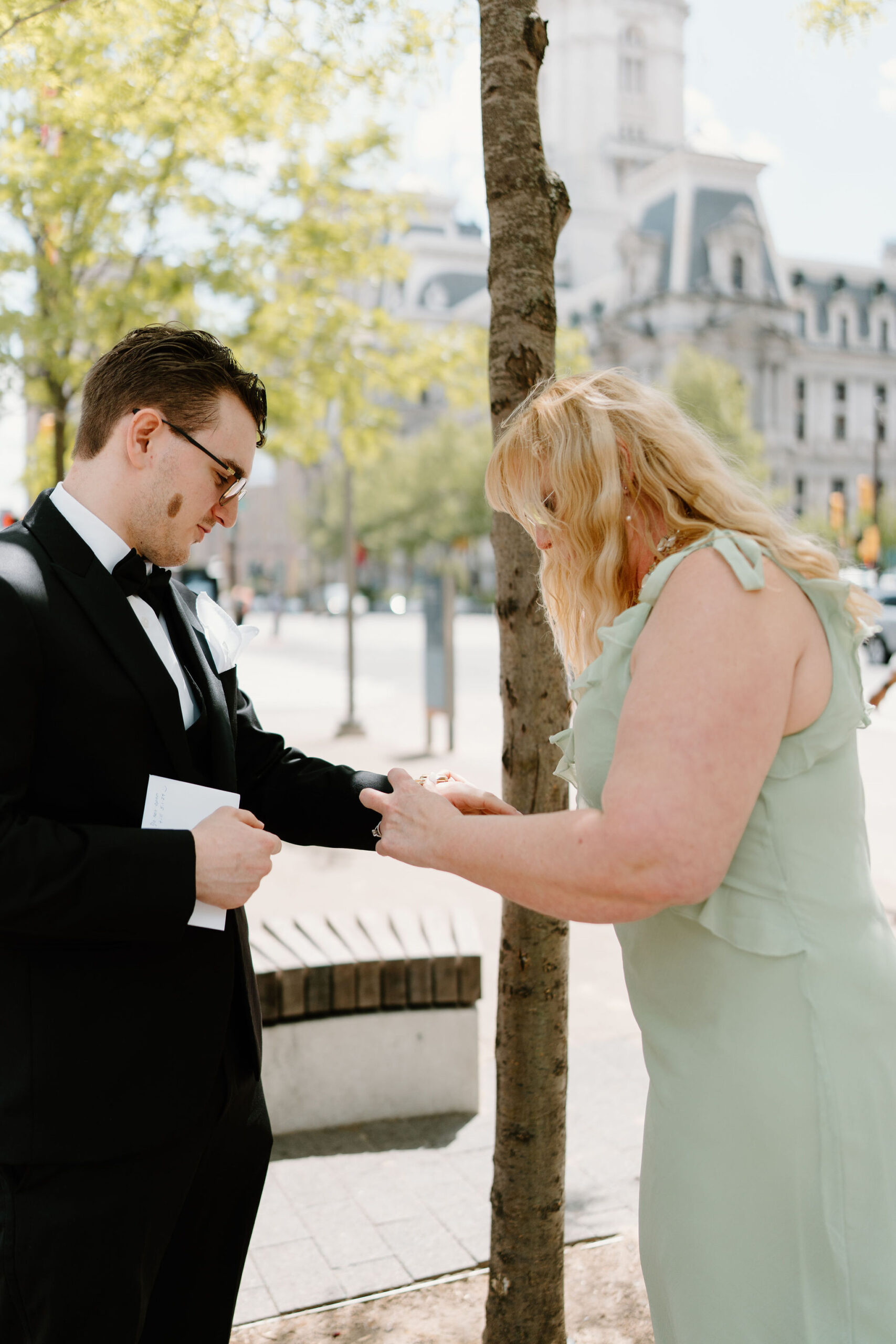 bride's mother (blonde hair, green dress) helping groom (black suit, glasses) to put on a watch