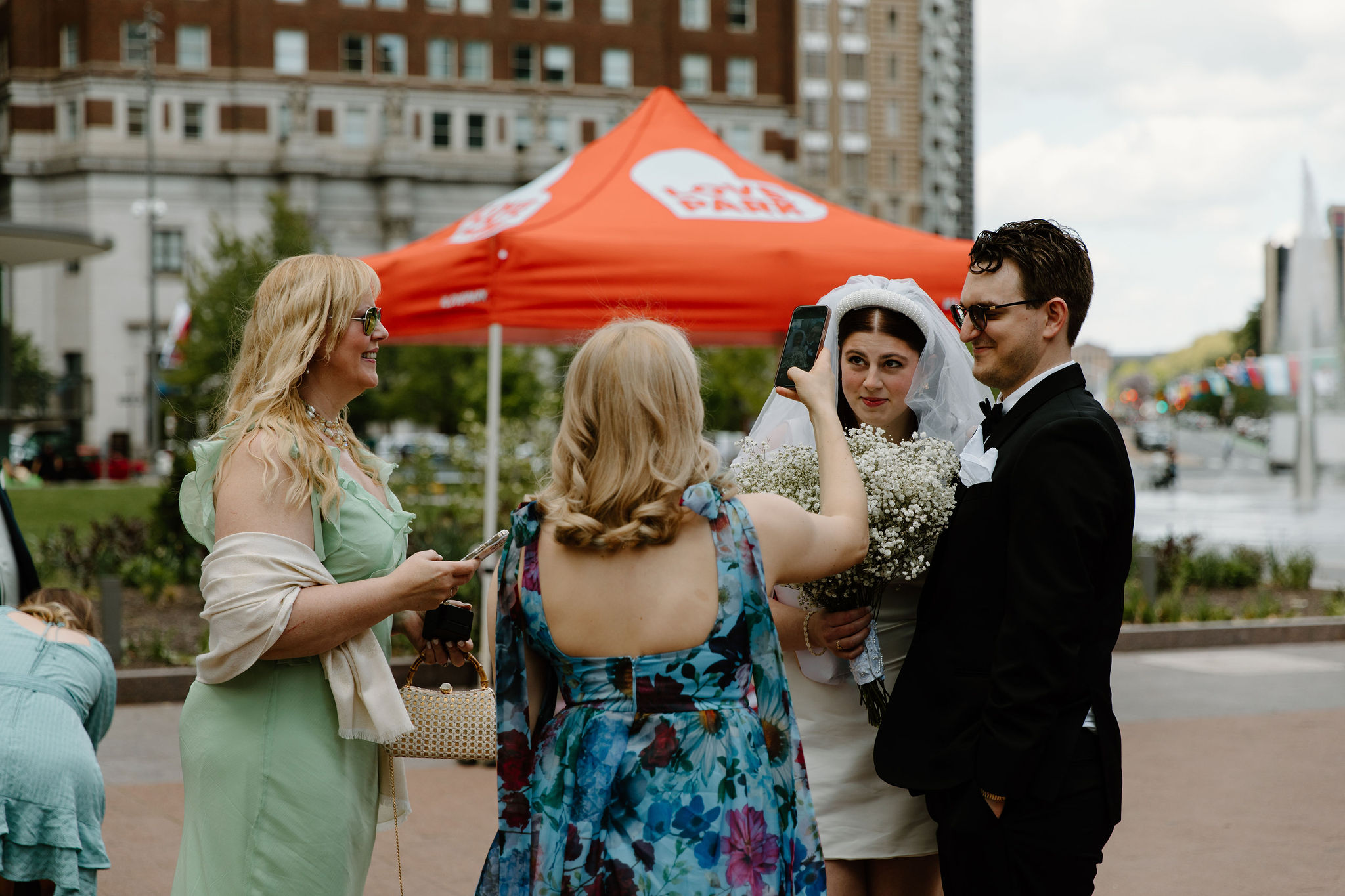 bride and groom posing, straight faced, while one of their sisters takes a 0.5 photo of them