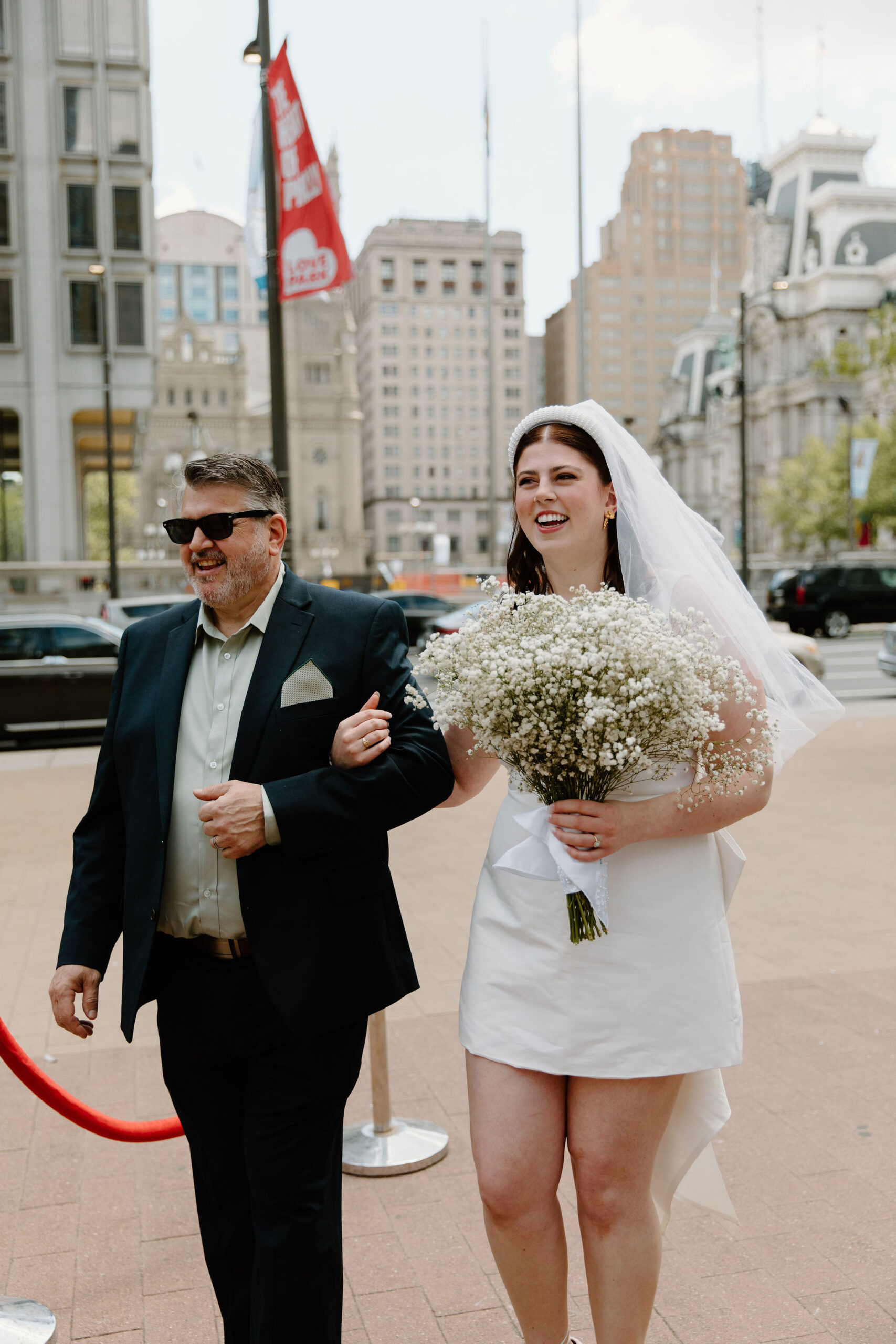 bride (short white dress, baby's breath bouquet, headband veil) and father (black suit) walking down the aisle for a wedding ceremony