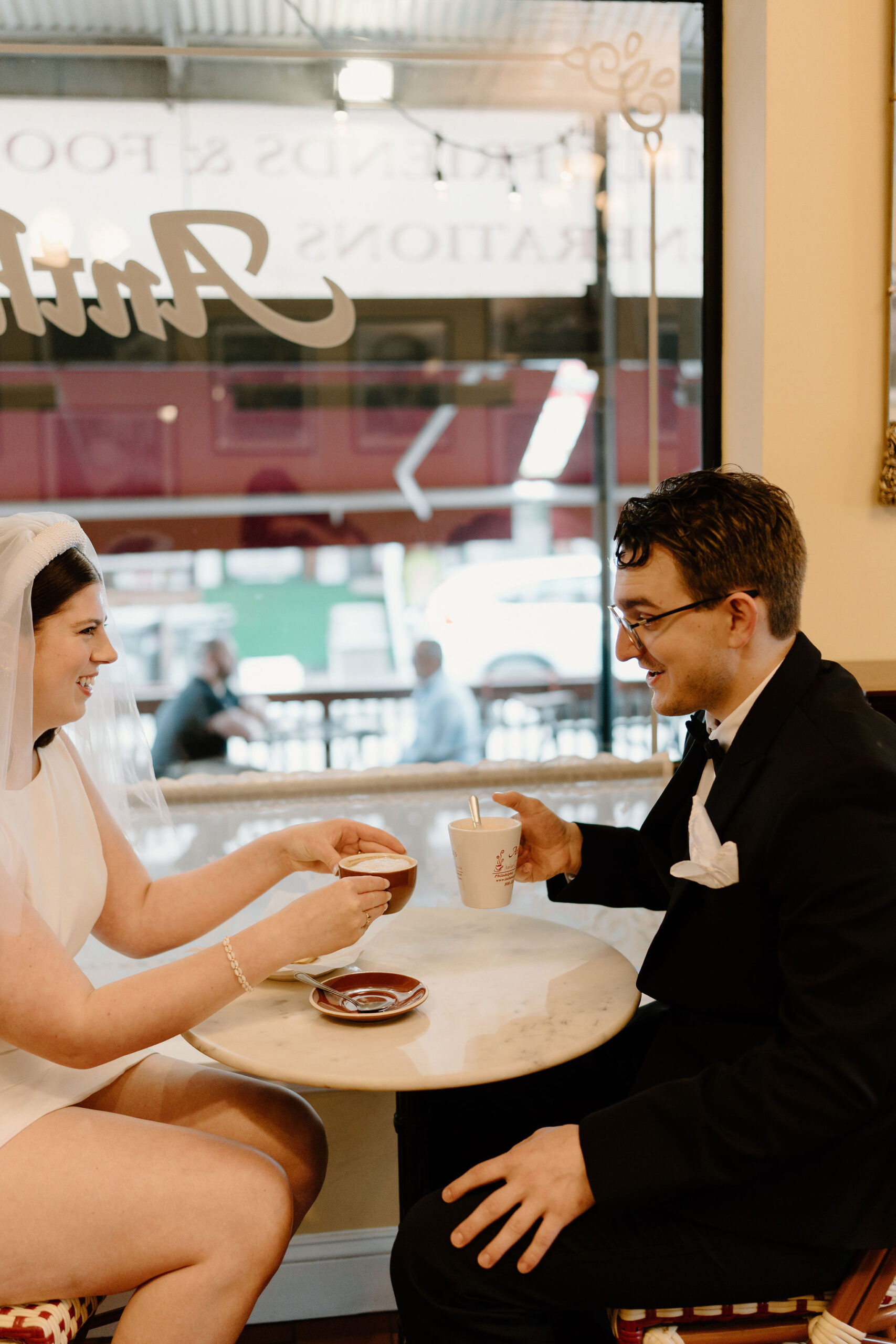 bride (short white dress, headband-veil) seated and toasting a coffee with groom (black suit, glasses) at a table by a window