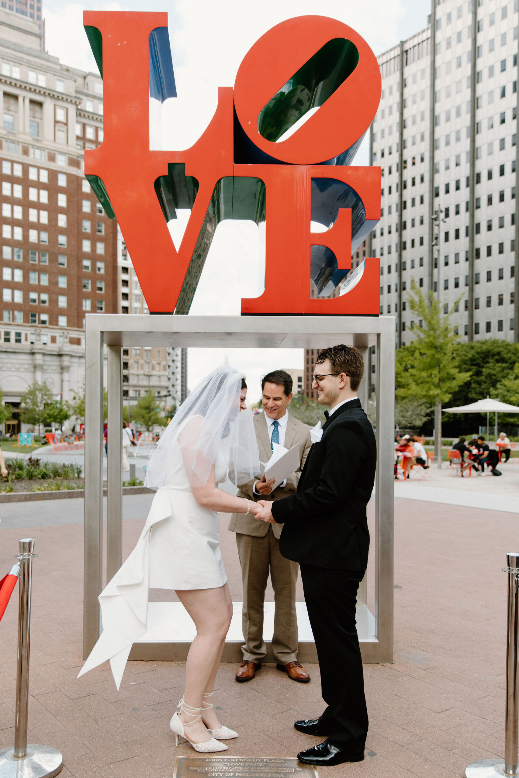 bride and groom laughing toward each other, standing with officiant in front of the Love Park sign