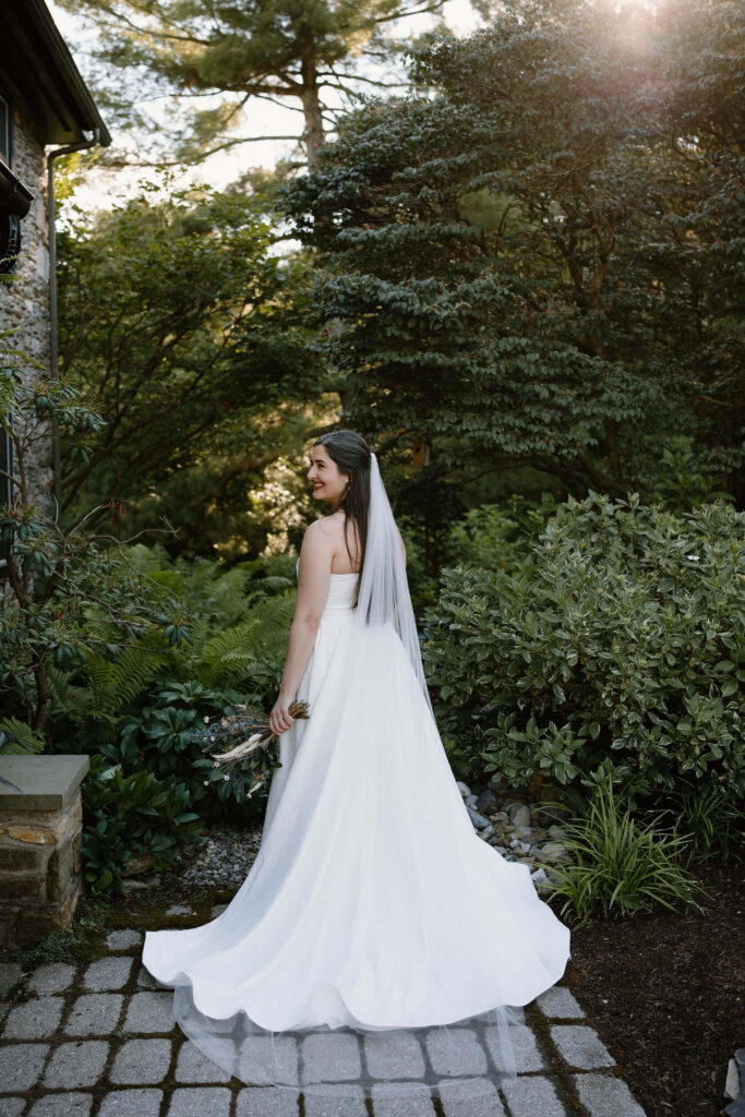 bride with a long white dress and long veil facing away from the camera, and looking over her shoulder off camera, holding flowers down at her side, with lots of trees and lush green bushes behind her