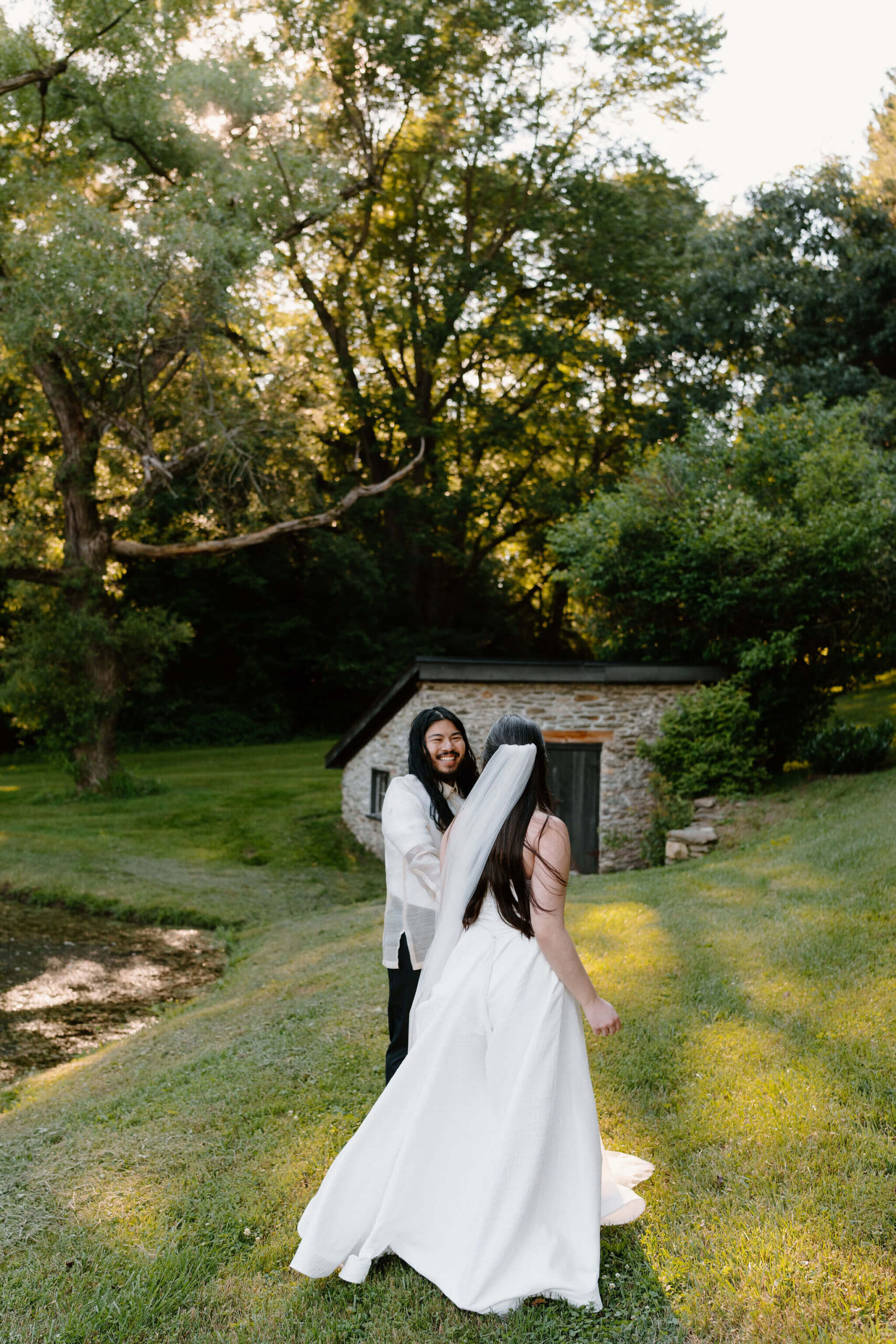 bride and groom next to a pond, surrounded by tall green trees, dancing and smiling at each other