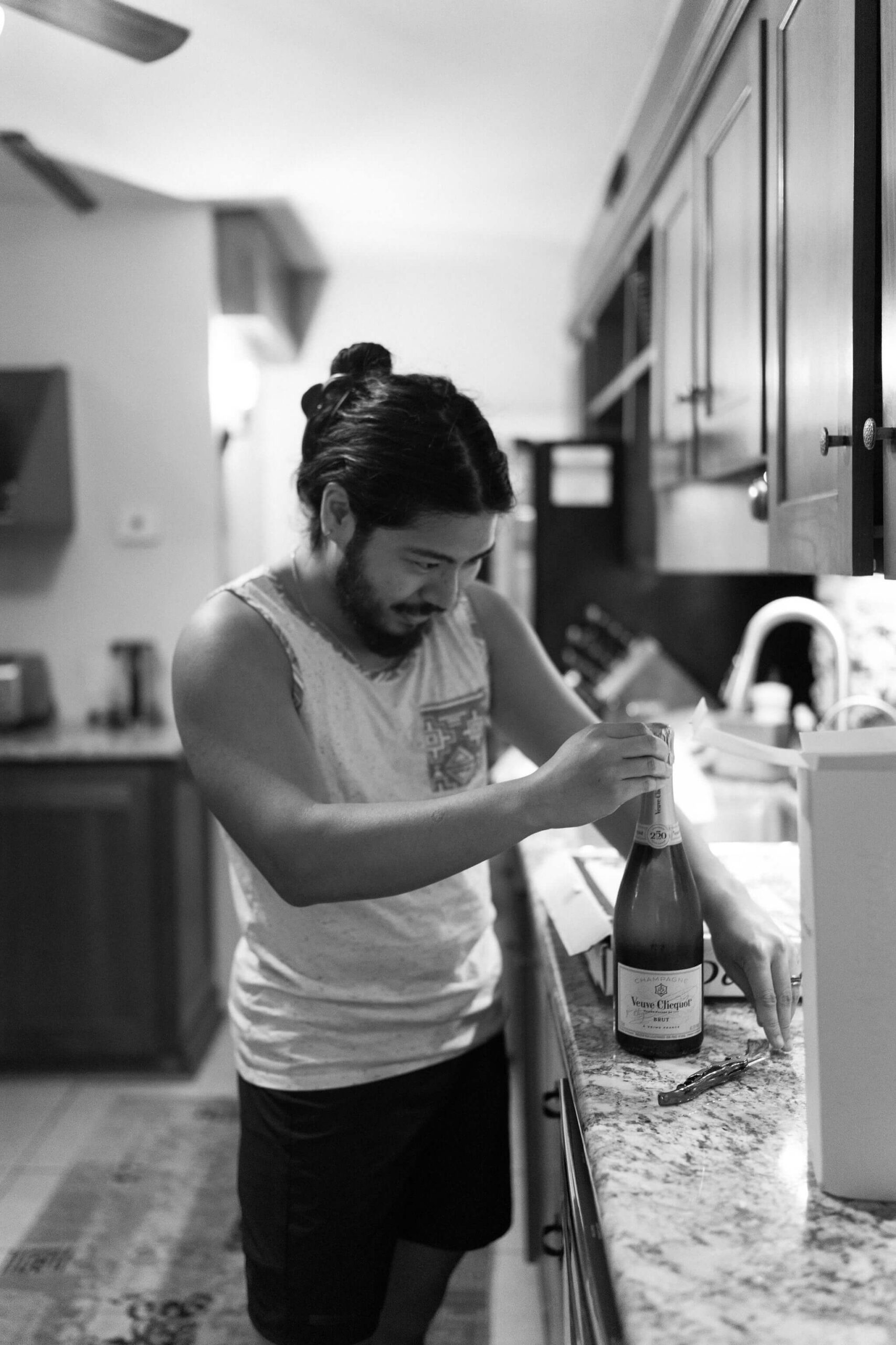 black and white image of groom, now wearing pajamas, holding a bottle of champagne, peeling off the foil to open it