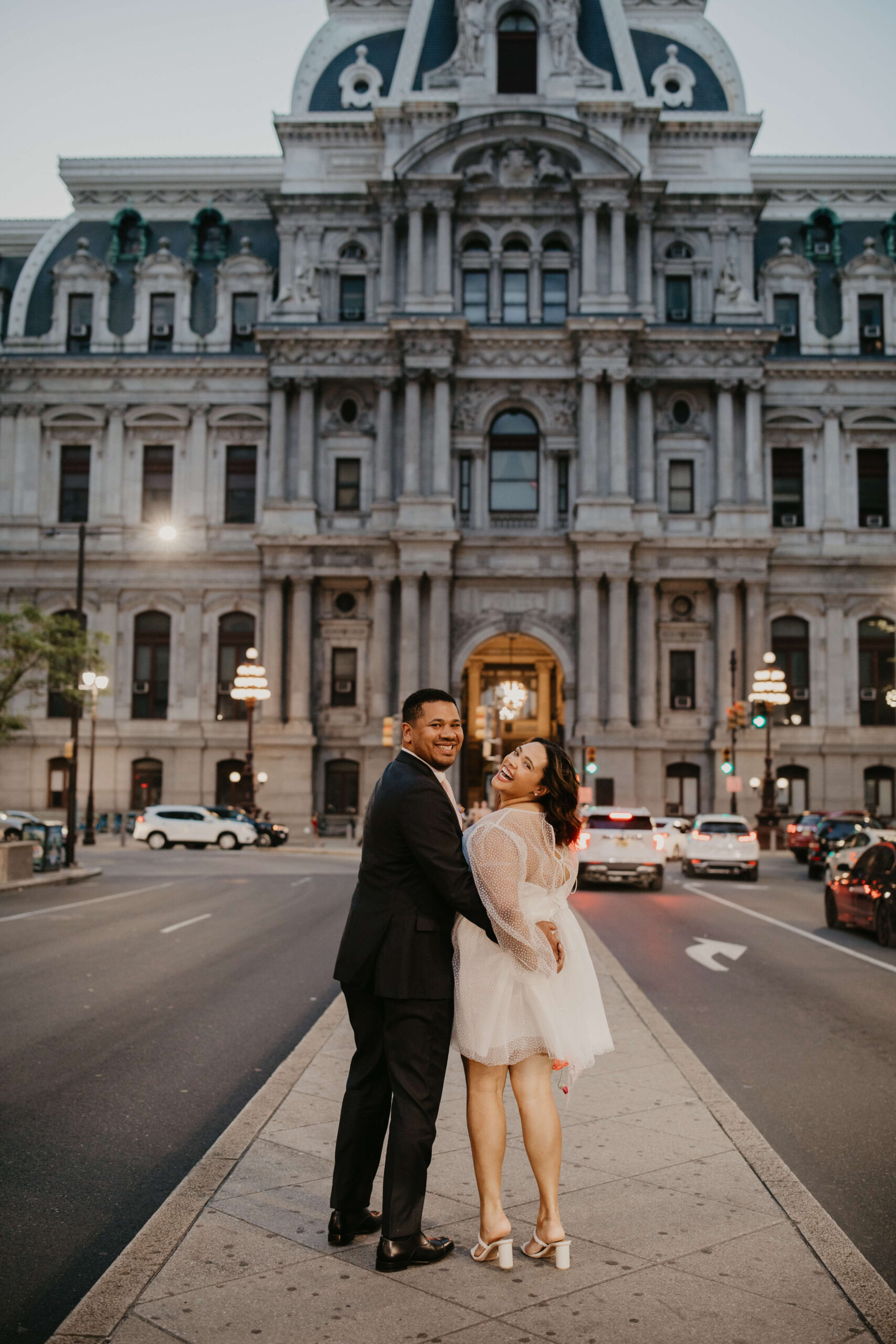 bride and groom walking up Broad Street toward City Hall in Philadelphia, looking back and smiling toward the camera while holding hands