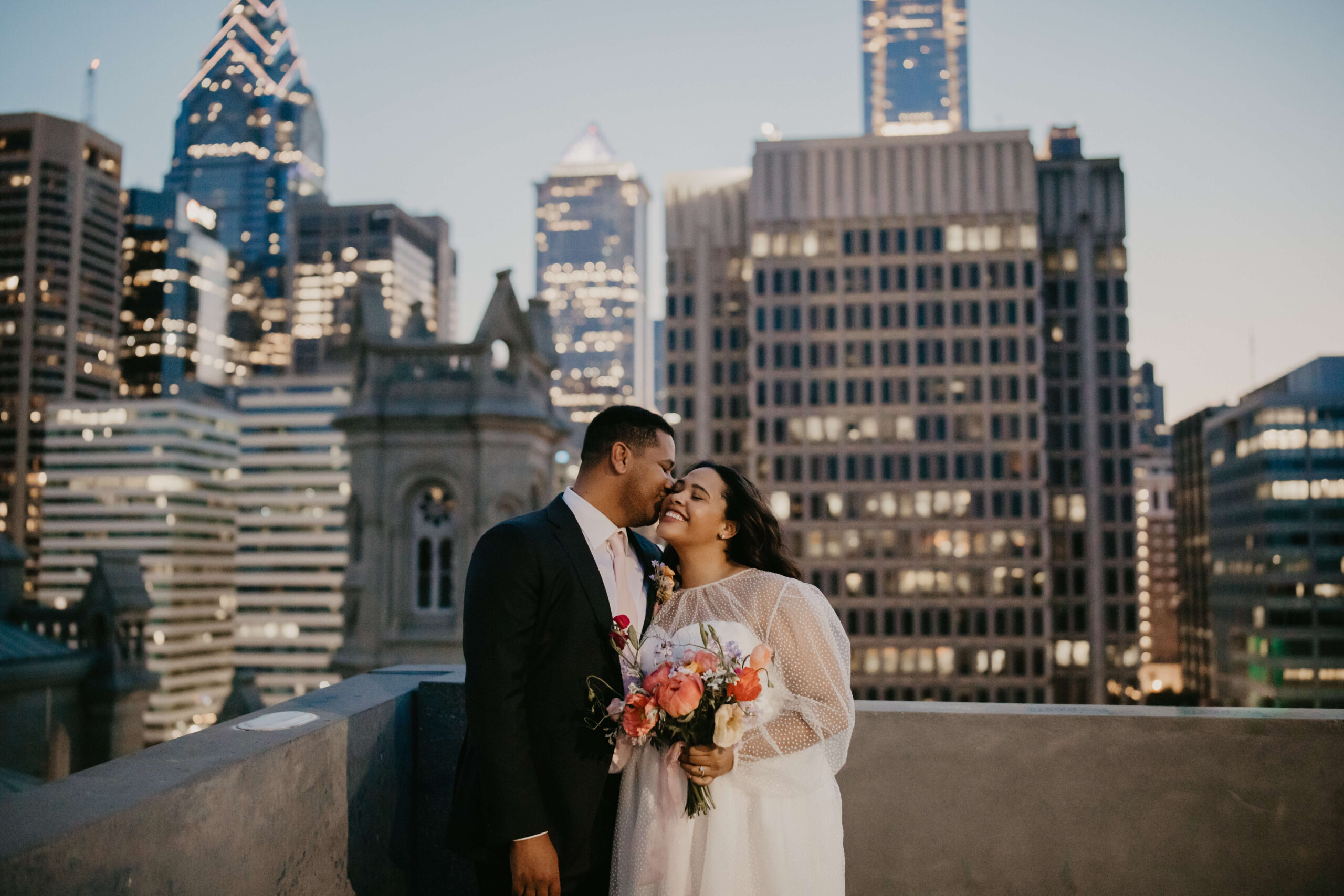 groom kissing bride on the cheek on top of a parking garage in center city Philadelphia, with the city skyline lit up behind them