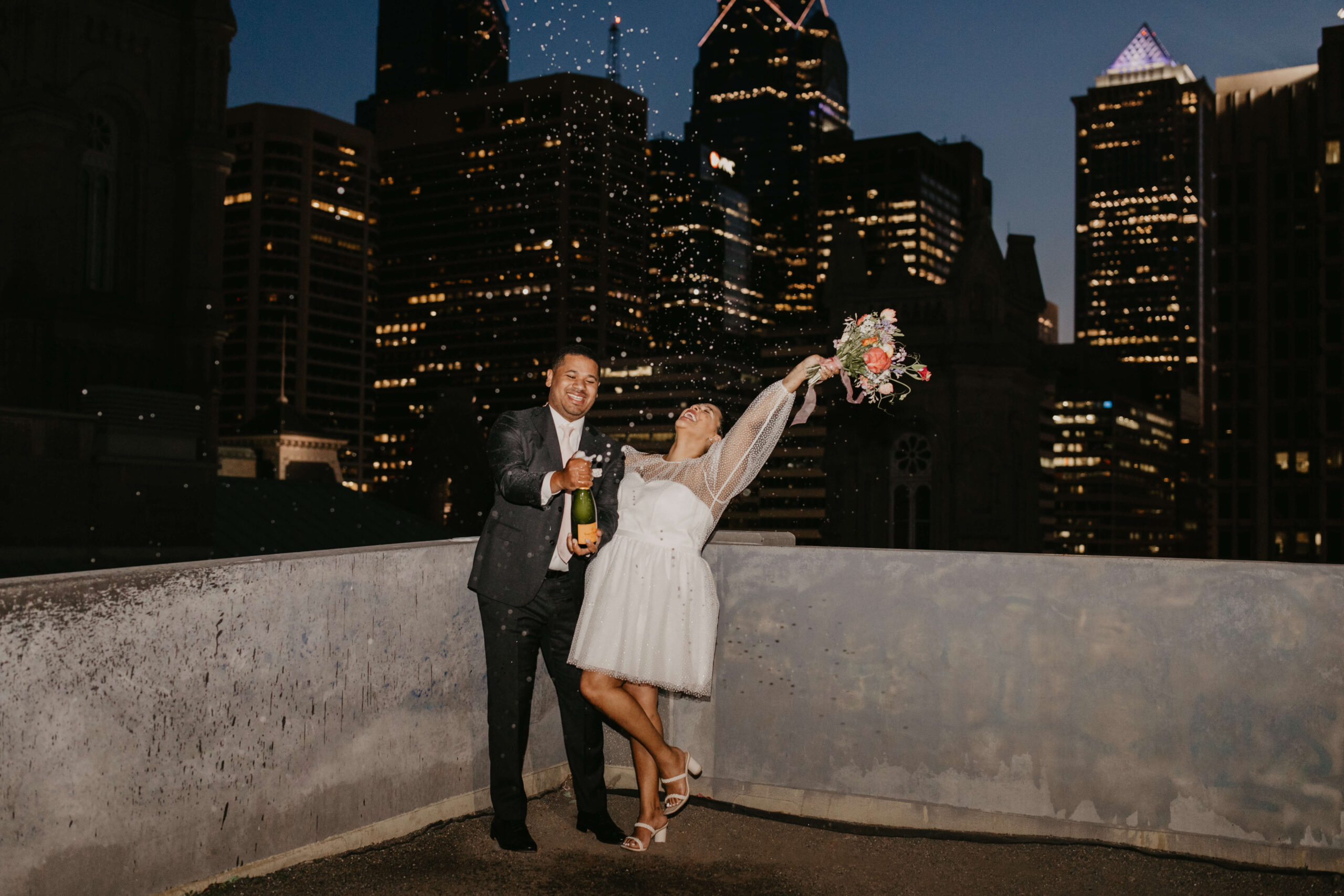 bride and groom celebrating and spraying champagne on top of a parking garage in center city Philadelphia, with the city skyline lit up behind them