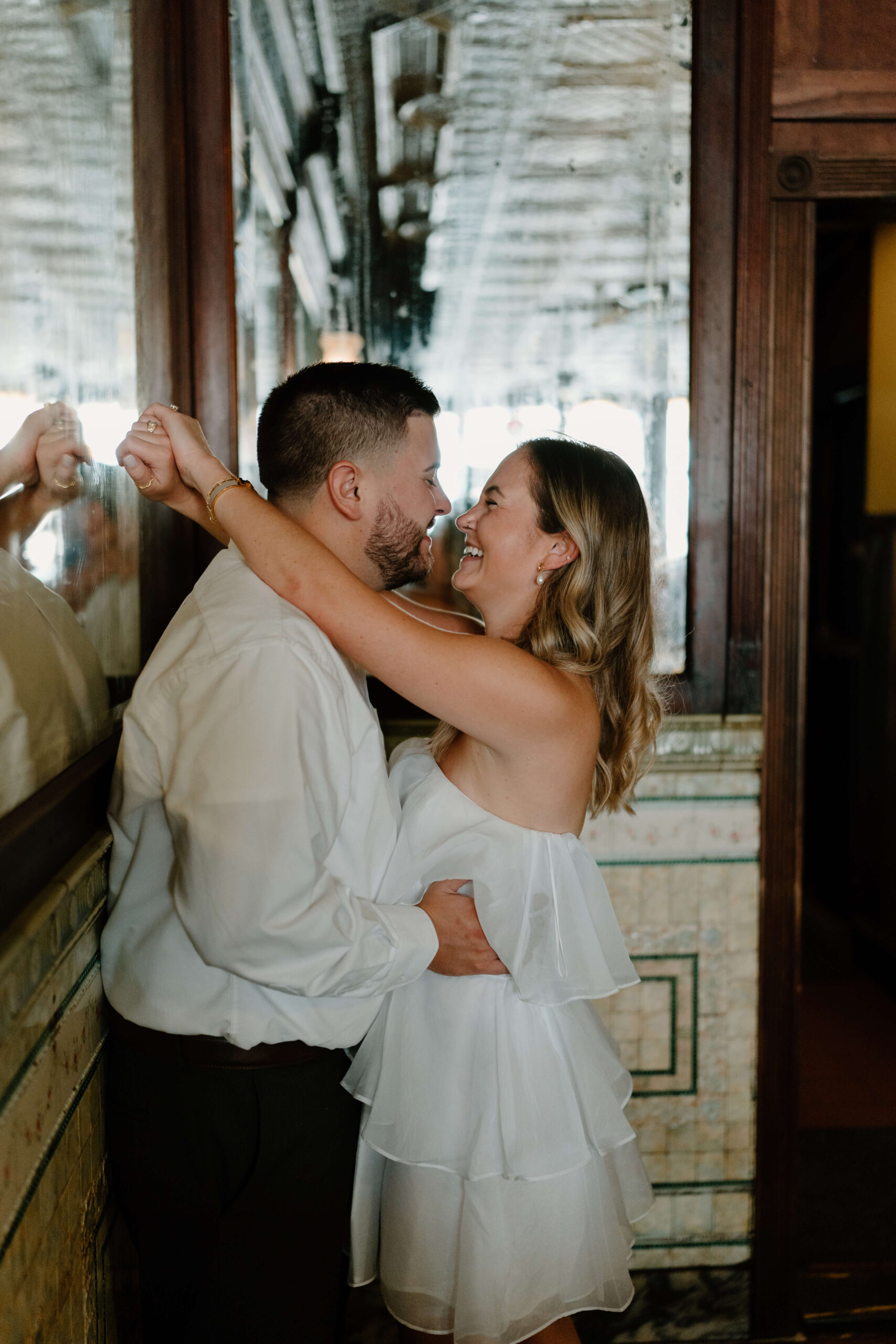 a couple embracing against a mirrored wall and smiling, both wearing white, with her arms thrown over his neck