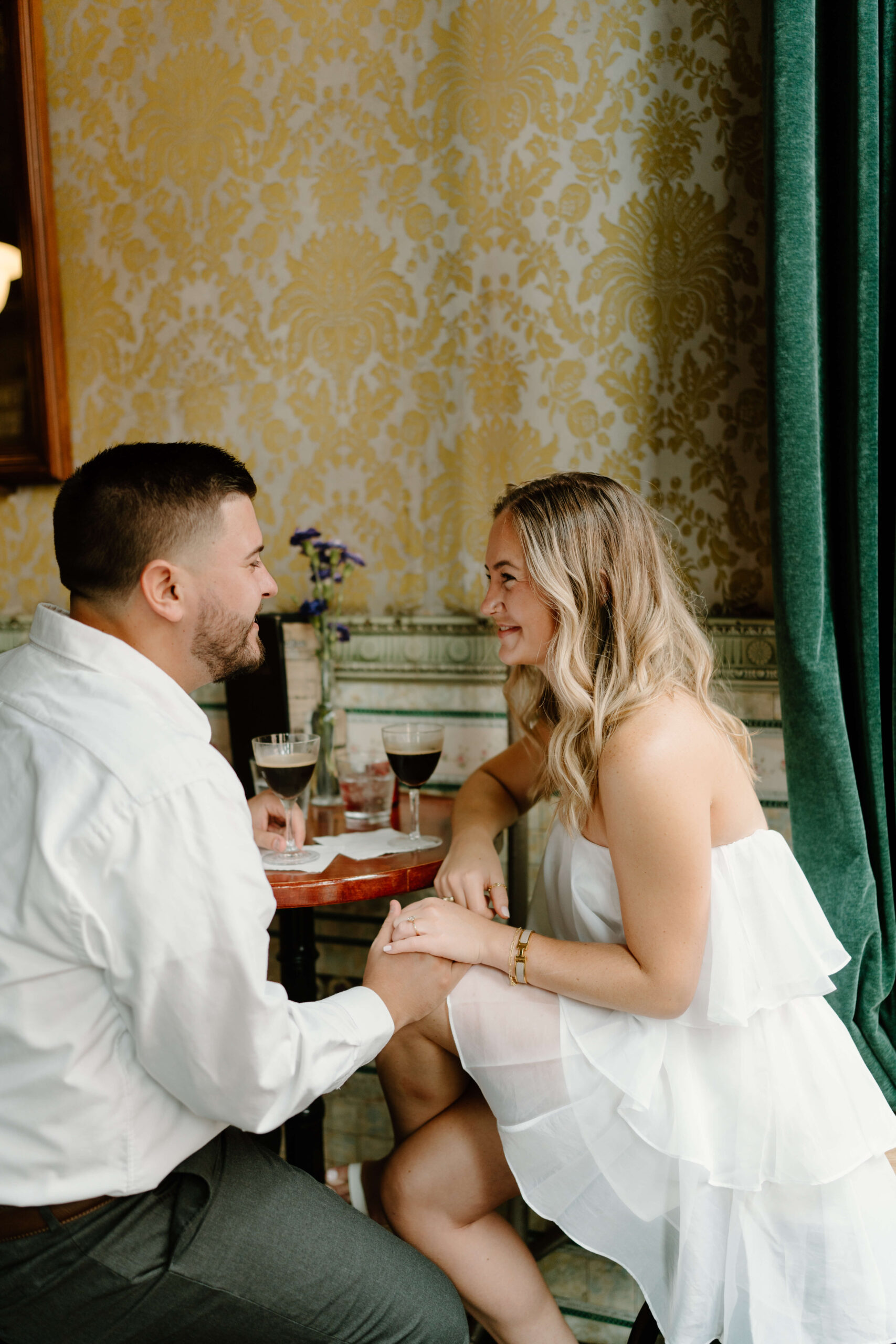 a couple, both wearing white, sitting at a table at a bar, both with espresso martinis, holding hands and smiling toward each other