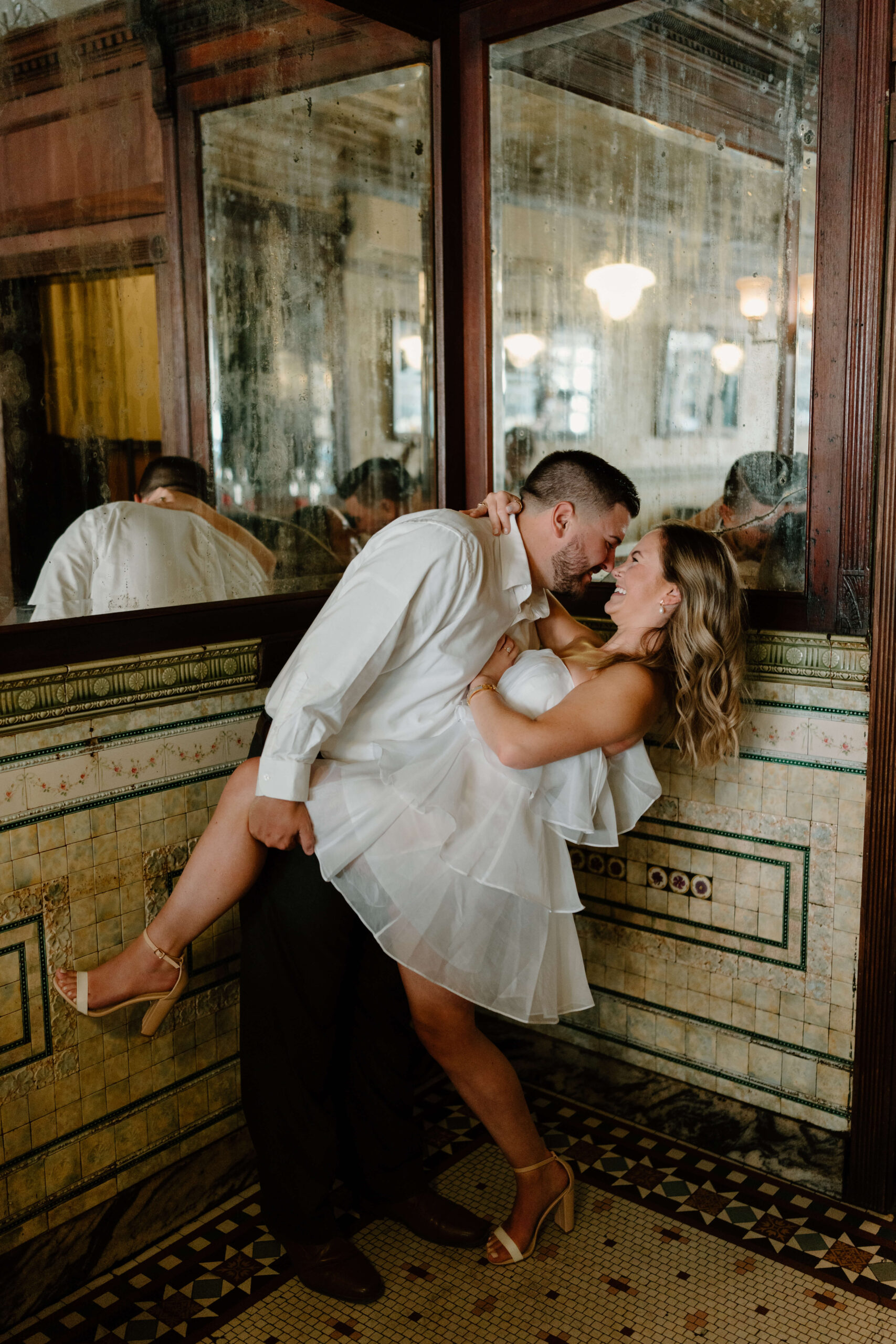 man dipping his fiance, both laughing, inside a restaurant with mirrored and tiled walls during their engagement pictures