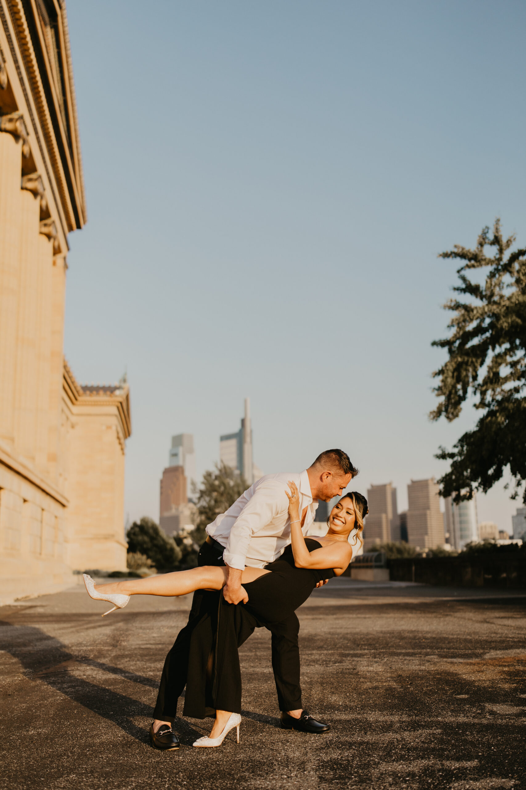 man dipping his wife, wearing a long black dress and heels, next to the Philadelphia Museum of Art, with the Philadelphia skyline behind them