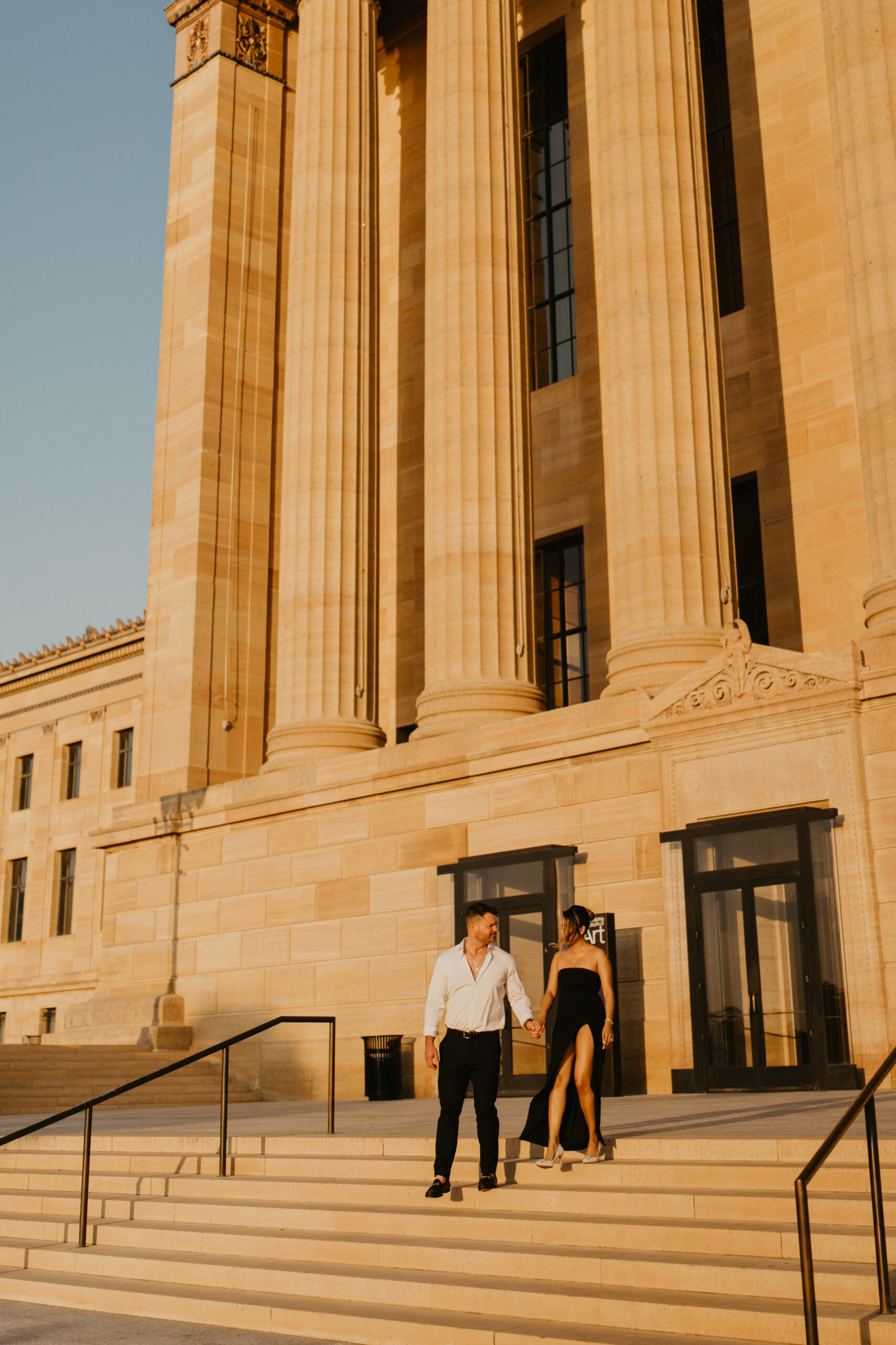 couples photos at the Philadelphia Museum of Art, featuring a man and a woman holding hands and walking down the art museum steps together, the pillars rising up behind them