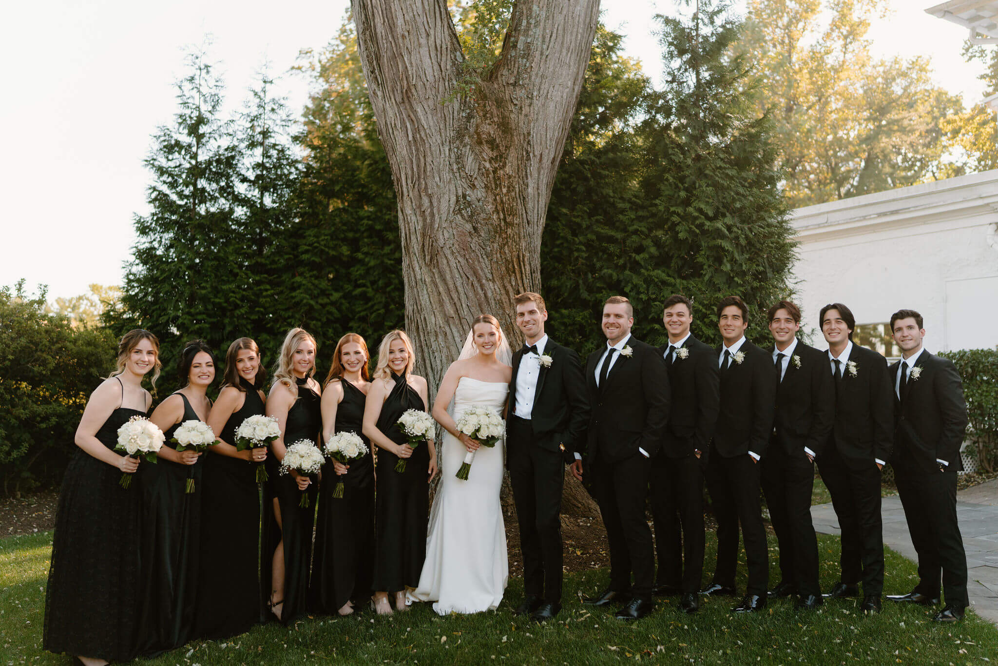 wedding party portrait with the bride and groom, all wearing black except for the bride, all smiling at the camera