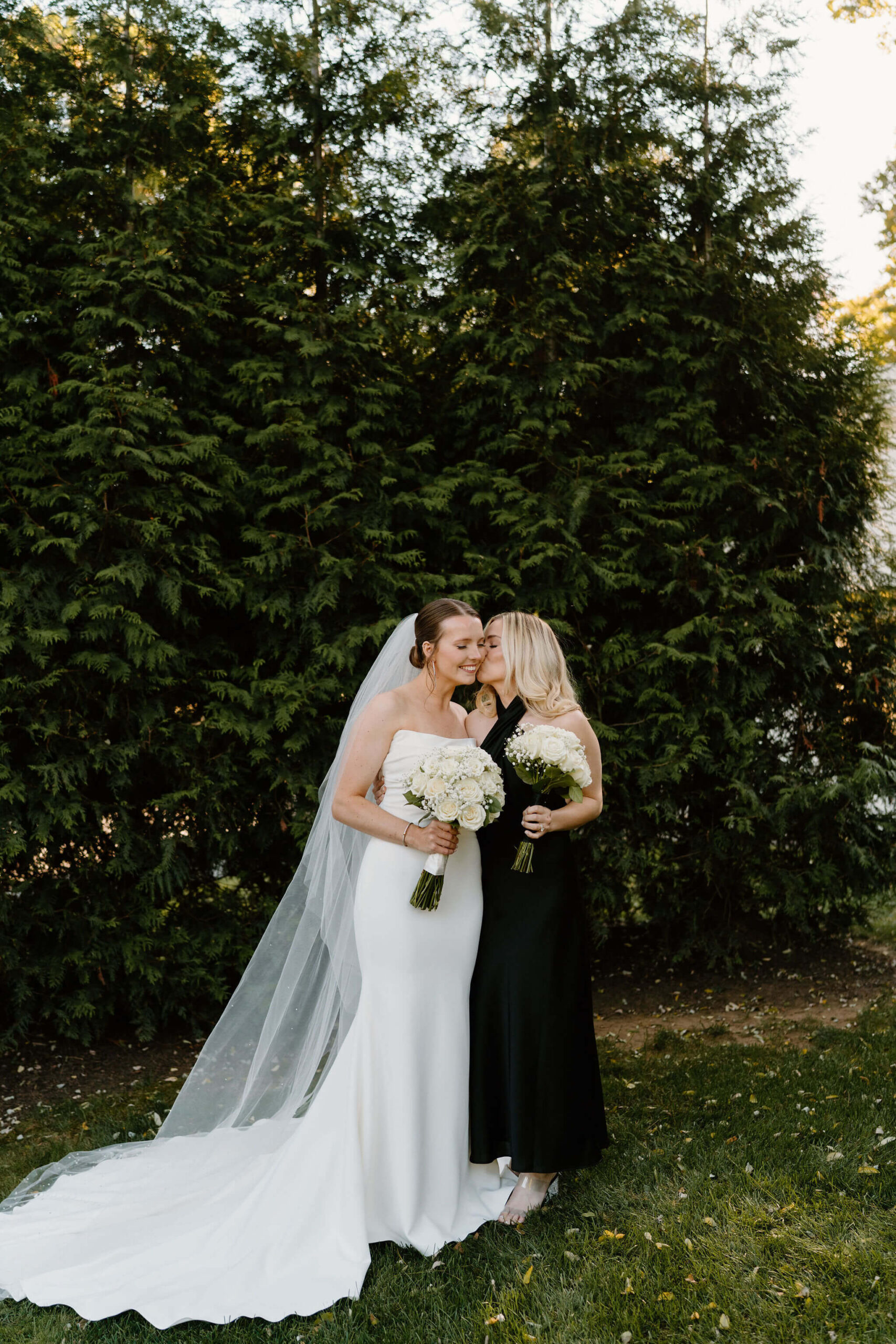 bride's sister and matron of honor wearing a black halter dress, kissing the bride on the cheek (wearing a white wedding gown, closing her eyes and smiling)