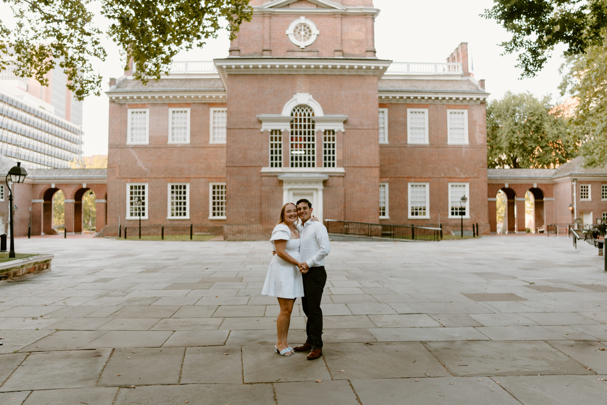 a couple smiling at the camera in front of independence hall in Washington Square Park in Philadelphia, PA for their engagement pictures