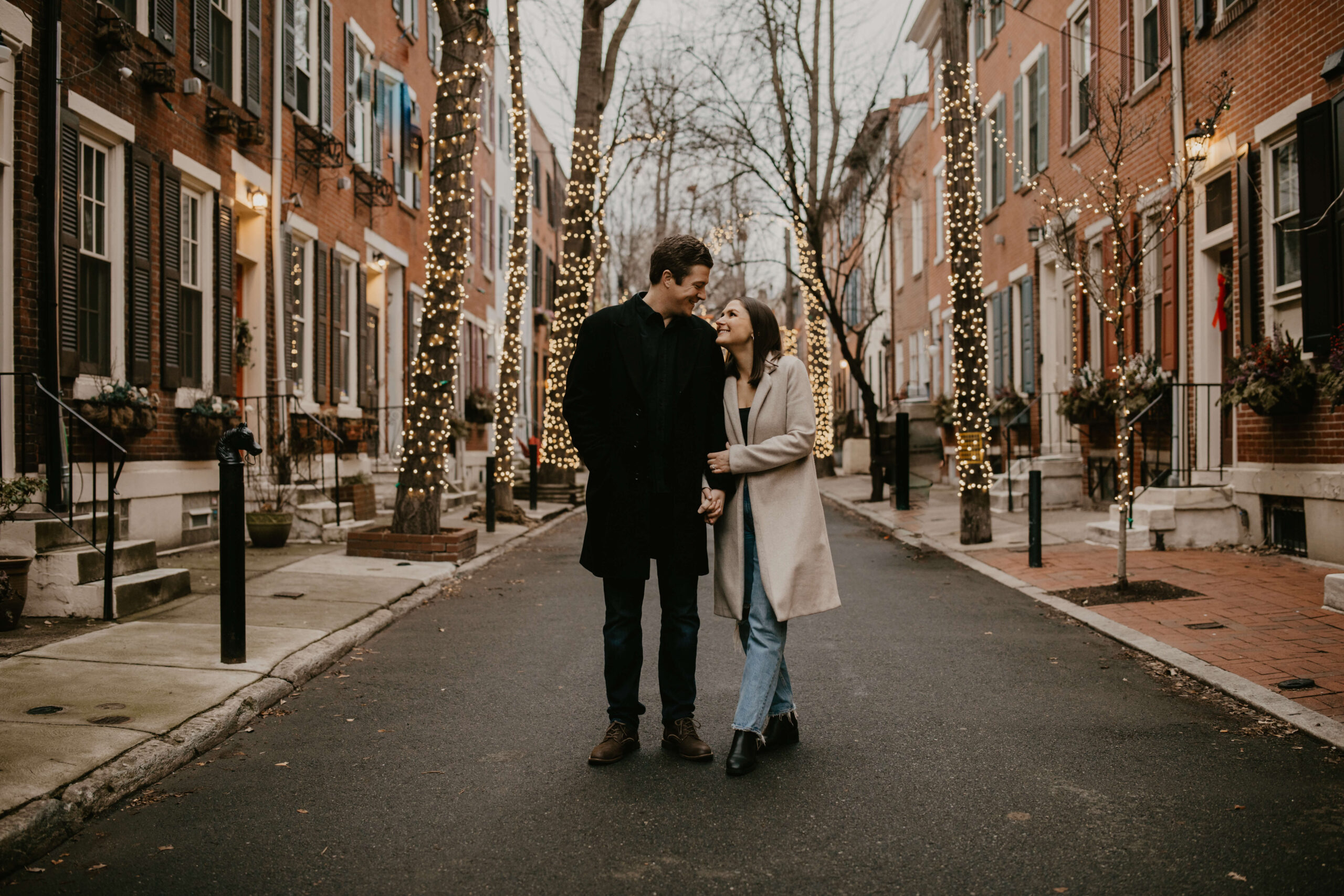 engaged couple holding hands and smiling at each other on Addison Street in RIttenhouse during their engagement pictures, old brick homes behind them and trees wrapped in white lights