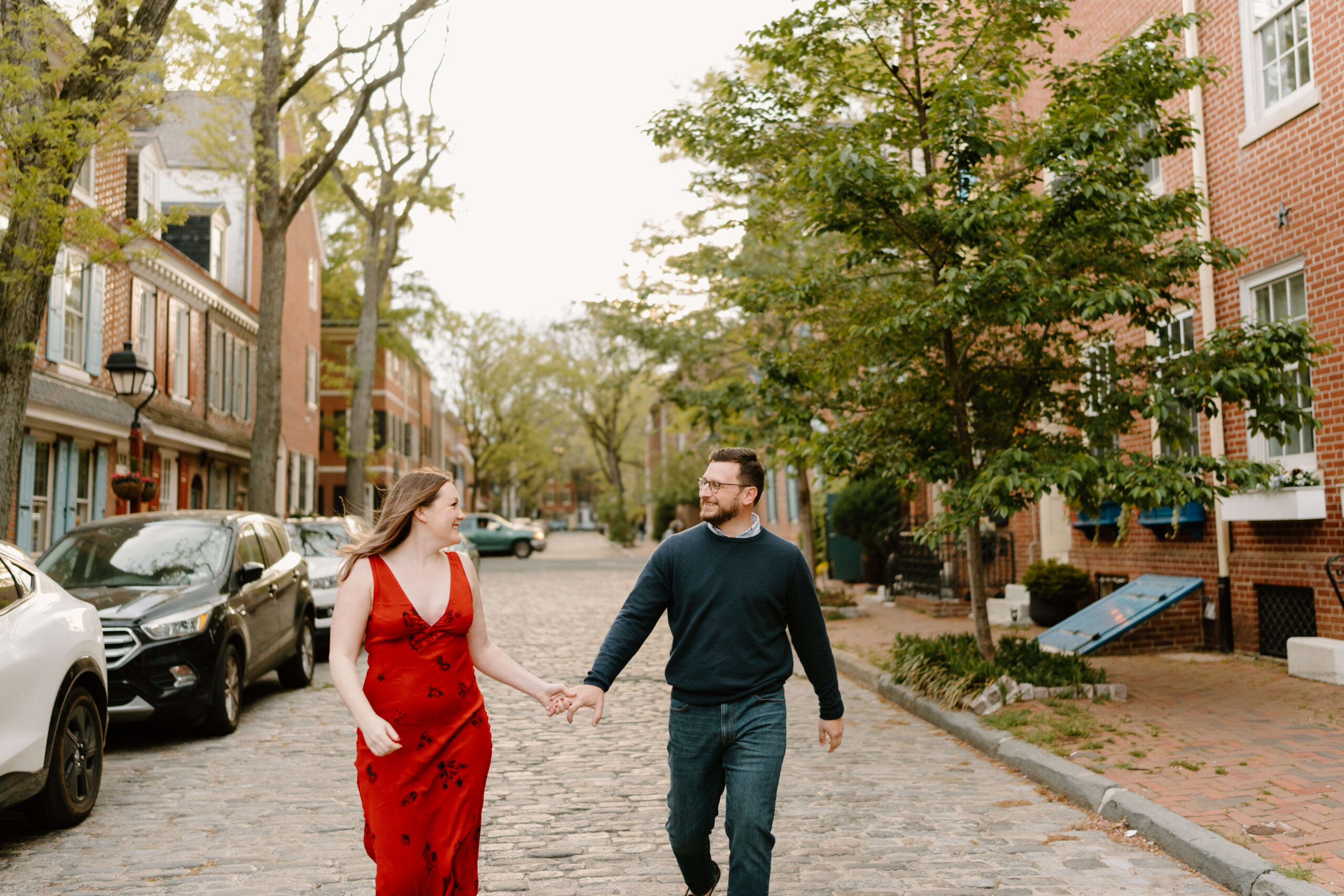 an engaged couple holding hands and walking down a cobblestone street, smiling at each other