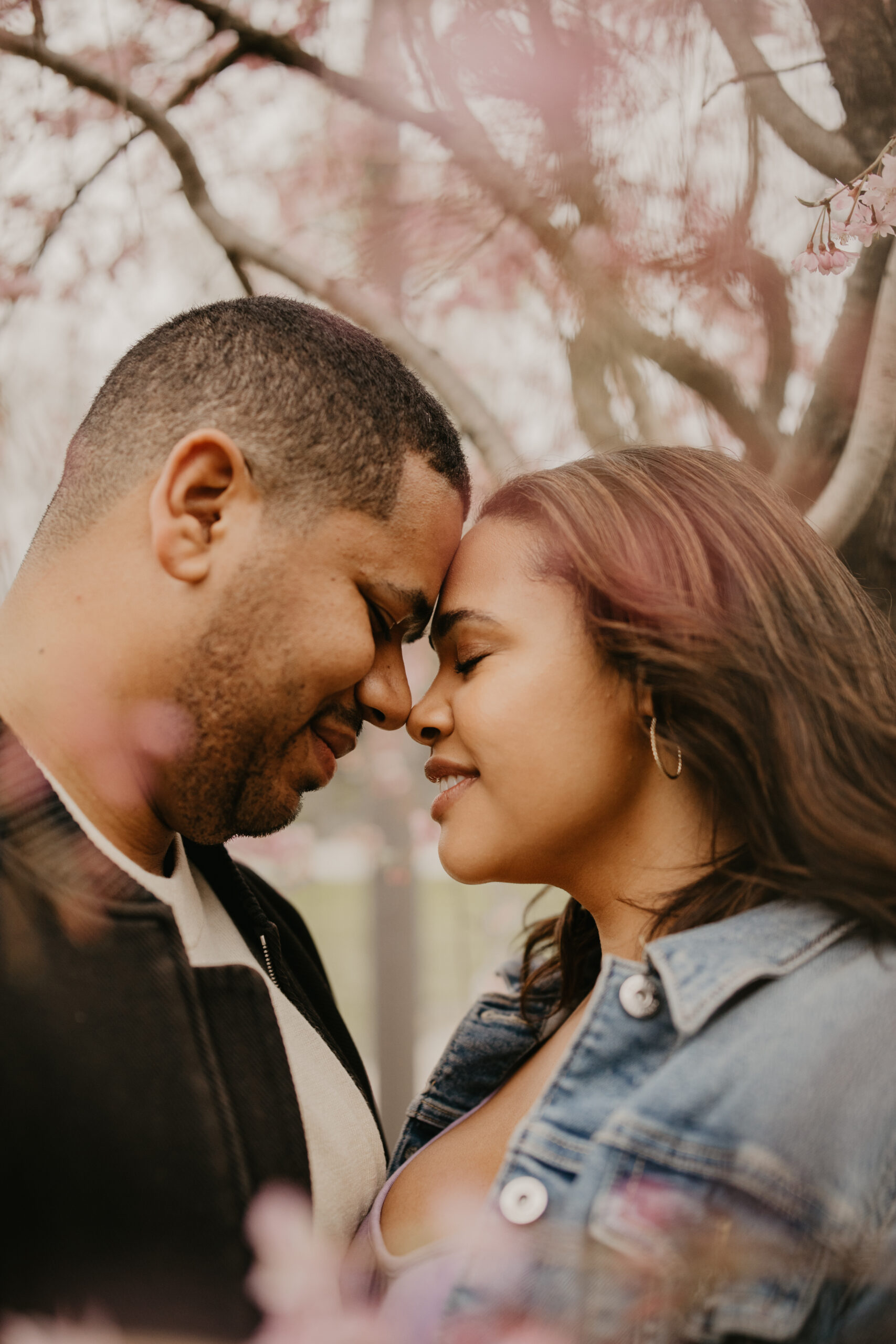 a couple standing nose-to-nose, with their eyes closed, underneath cherry blossoms