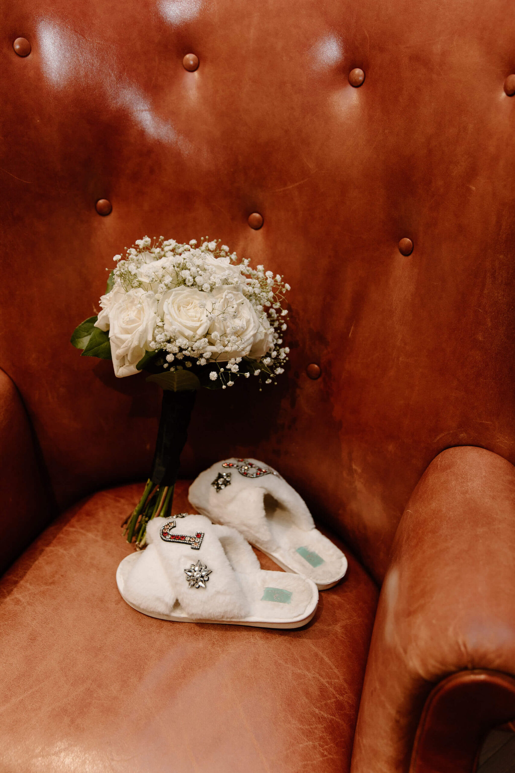 white bedazzled slippers and a bridesmaid's bouquet set out on a leather chair