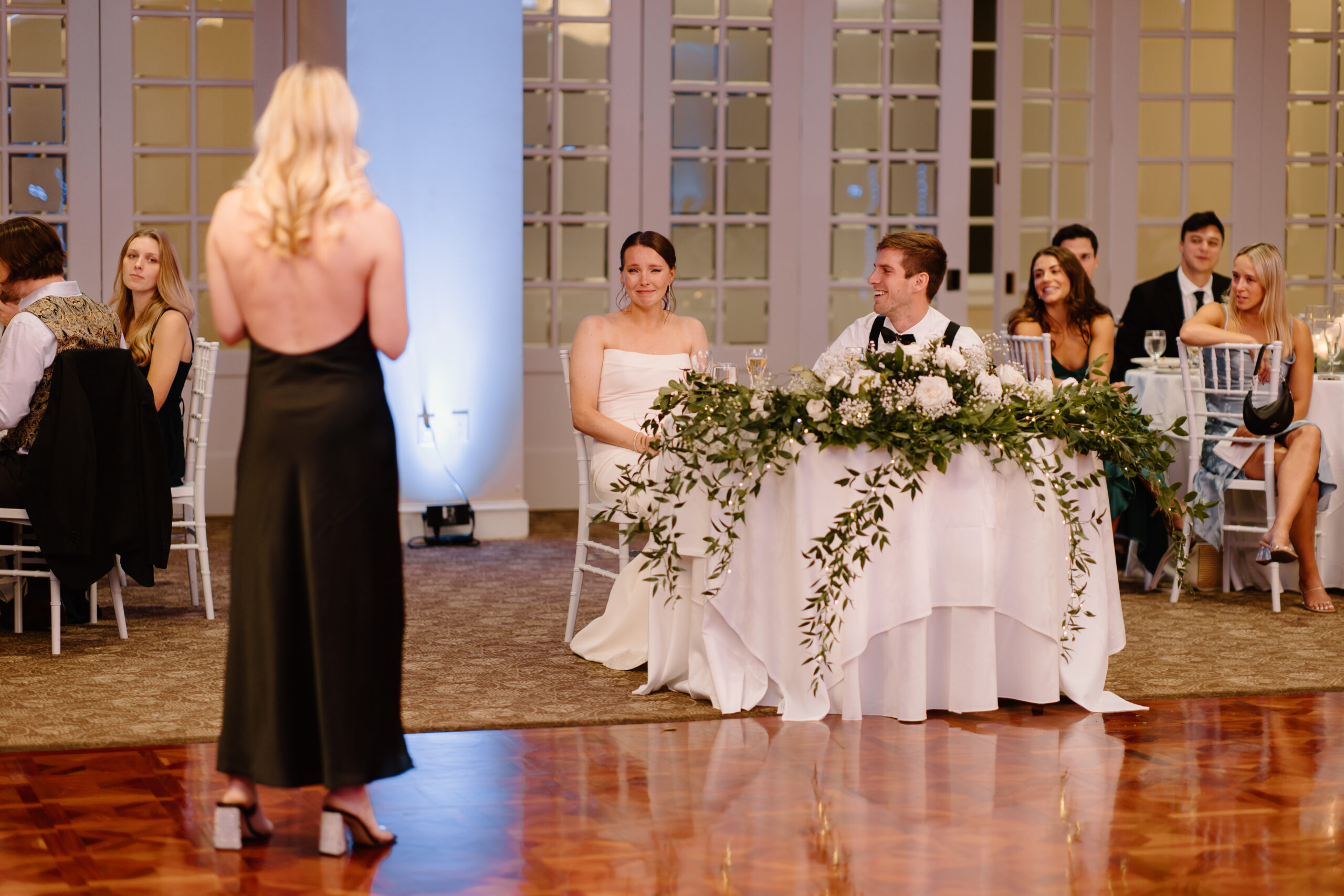 matron of honor giving speech at a wedding reception, while bride cries at the sweetheart table with her new husband