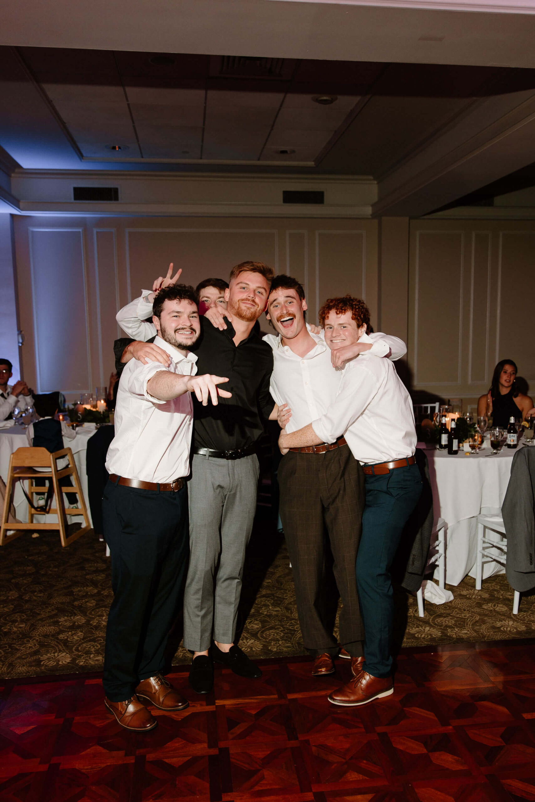 group of friends posing together on the dance floor at a wedding reception, arms around each other and smiling with one giving "bunny ears"