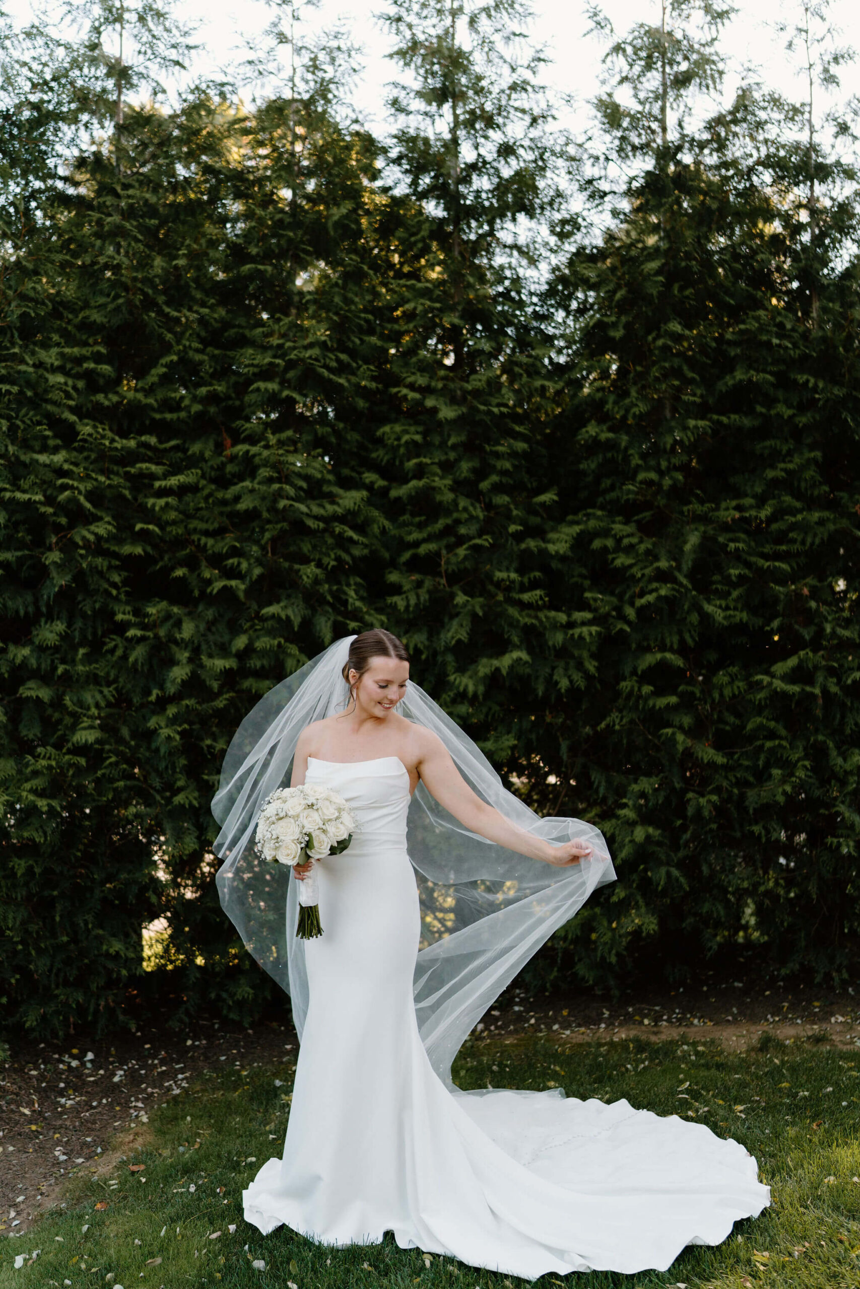 bride standing in front of large evergreen trees holding her bouquet and swinging her veil in her other hand
