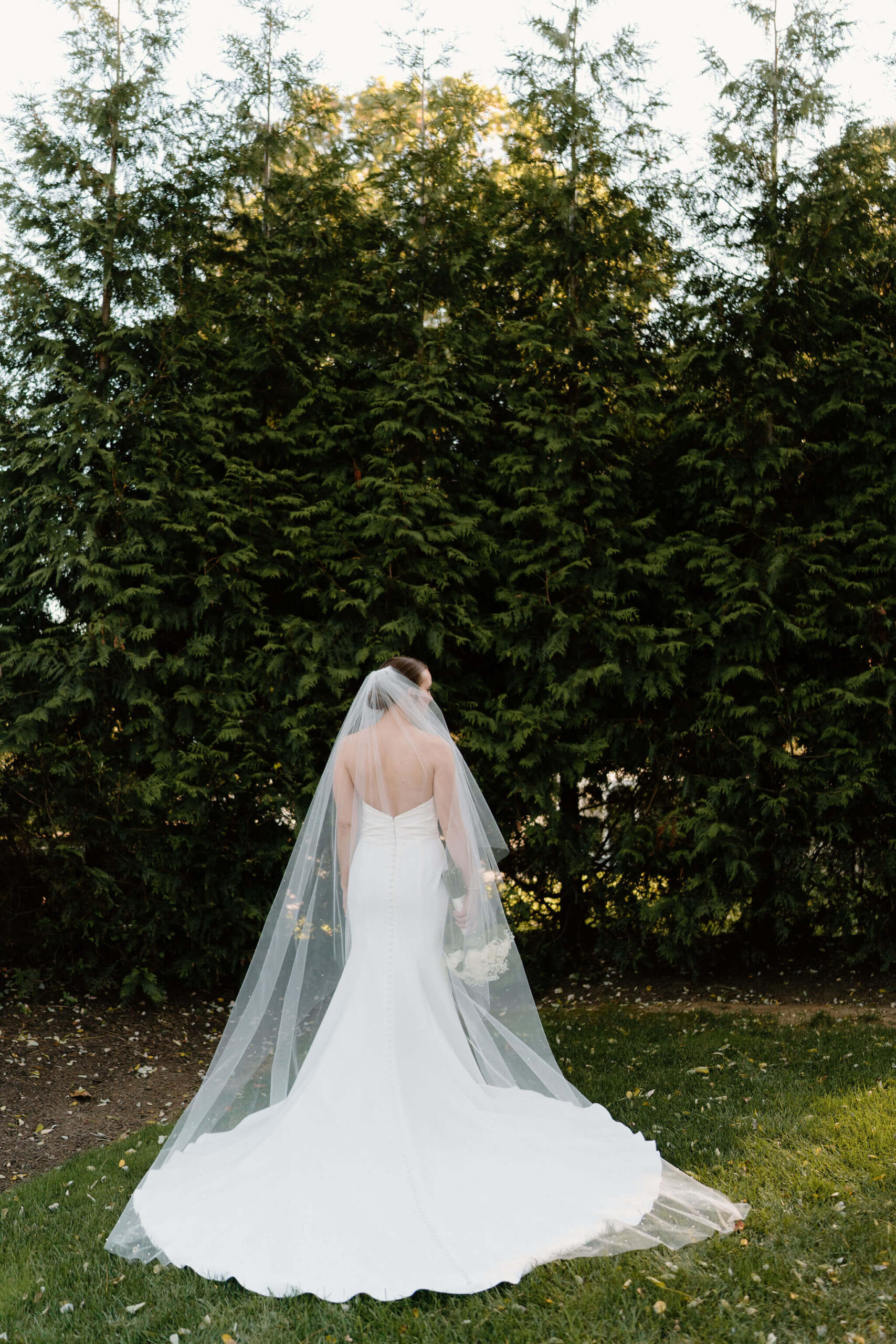 bride in a long white backless wedding dress and a long veil, facing away from the camera with her bouquet held down at her side