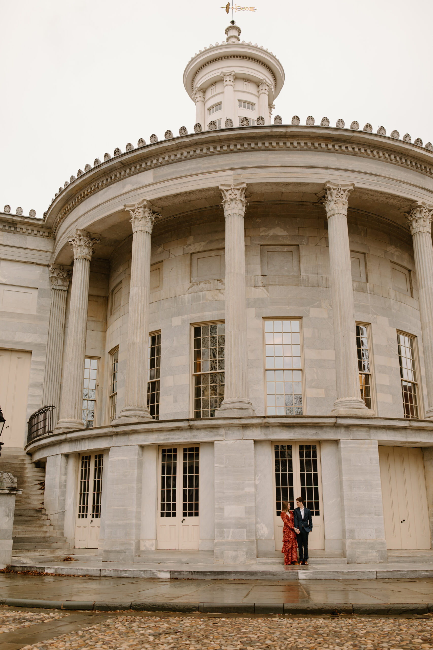 a couple (woman in a red dress, man in a navy blue suit) holding hands in front of the Merchant's Exchange Building in Philadelphia