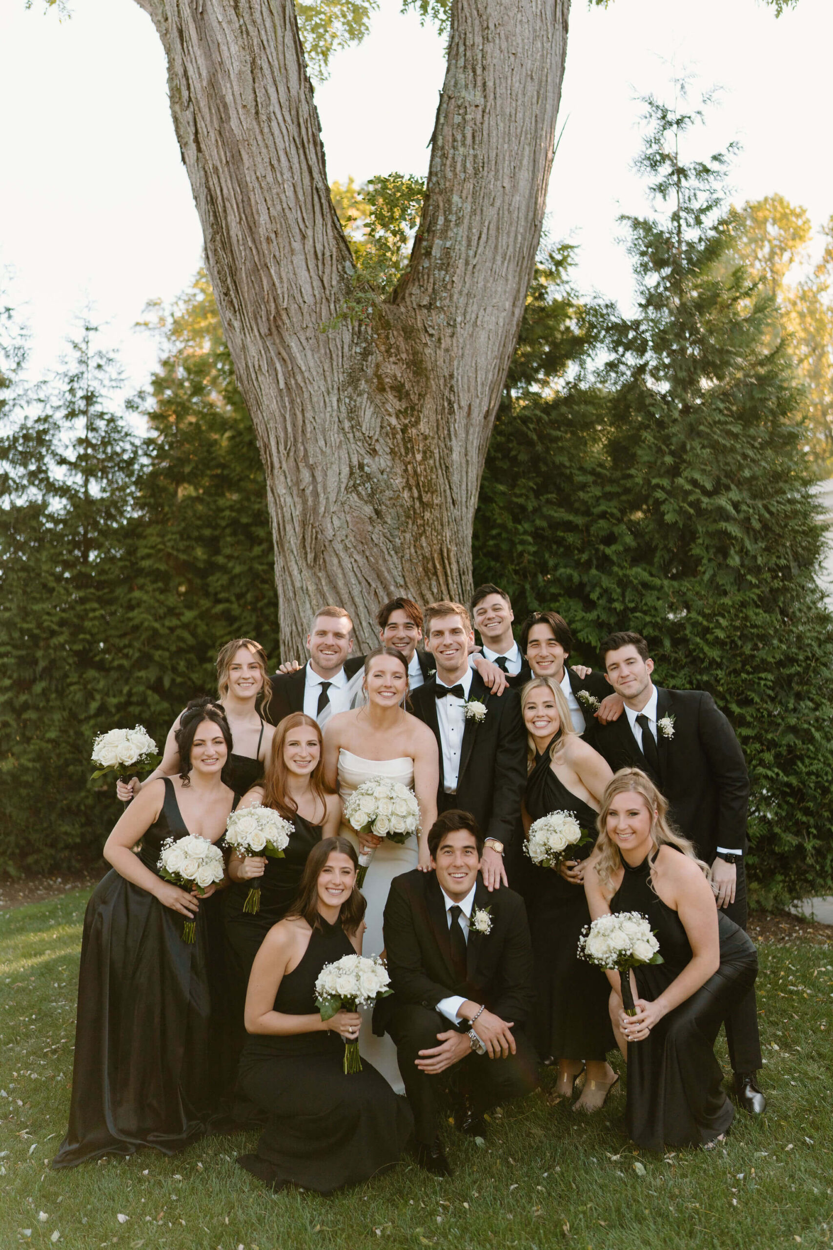 bride and groom (white wedding dress, black suit, respectively) surrounded by their wedding party (all wearing black dresses and suits with white roses), smiling at the camera