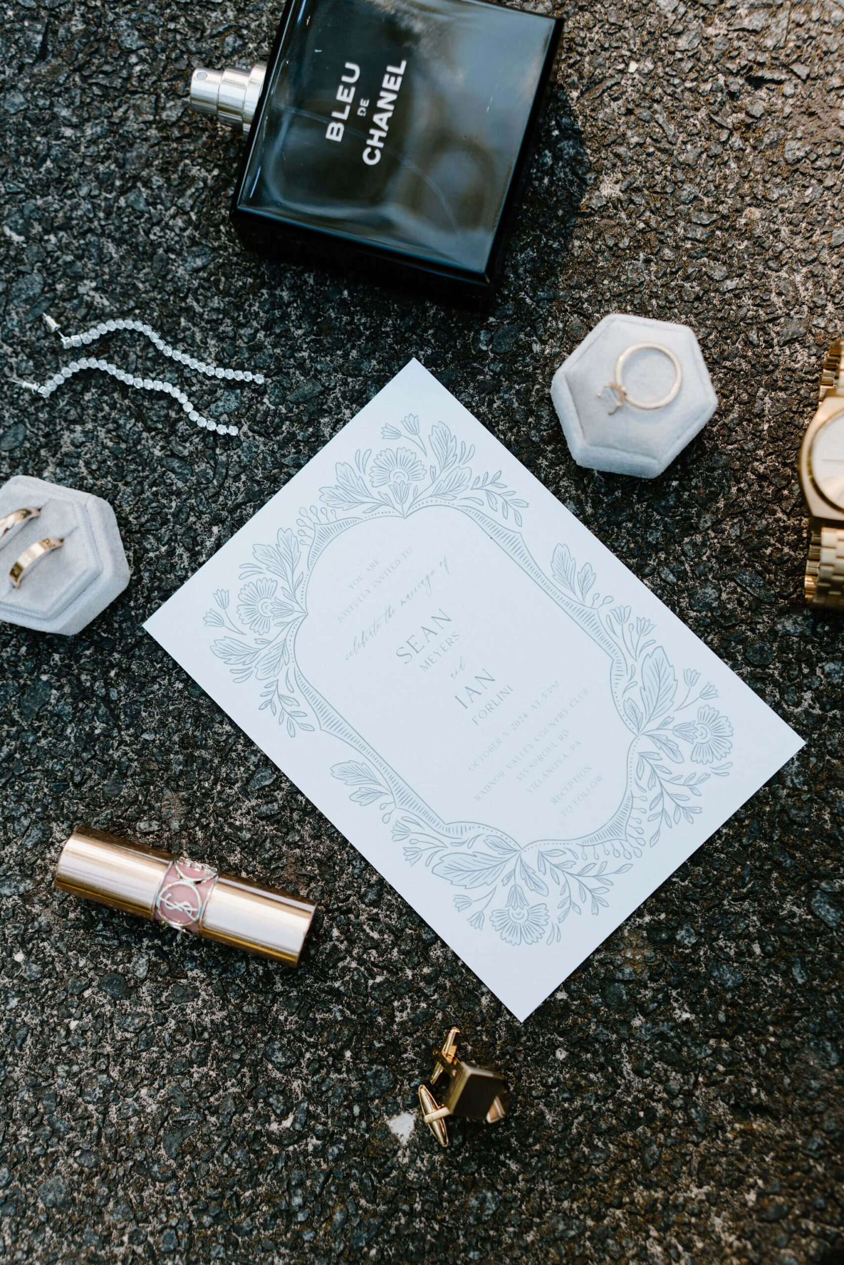 flat lay of a wedding invitation, engagement ring, wedding bands, lipstick, cuff links, watch, and cologne