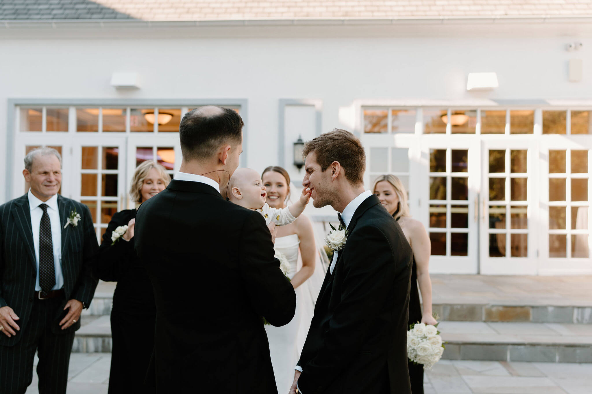 bride and groom's nephew reaching up and touching the groom's face while smiling, surrounding family members laughing