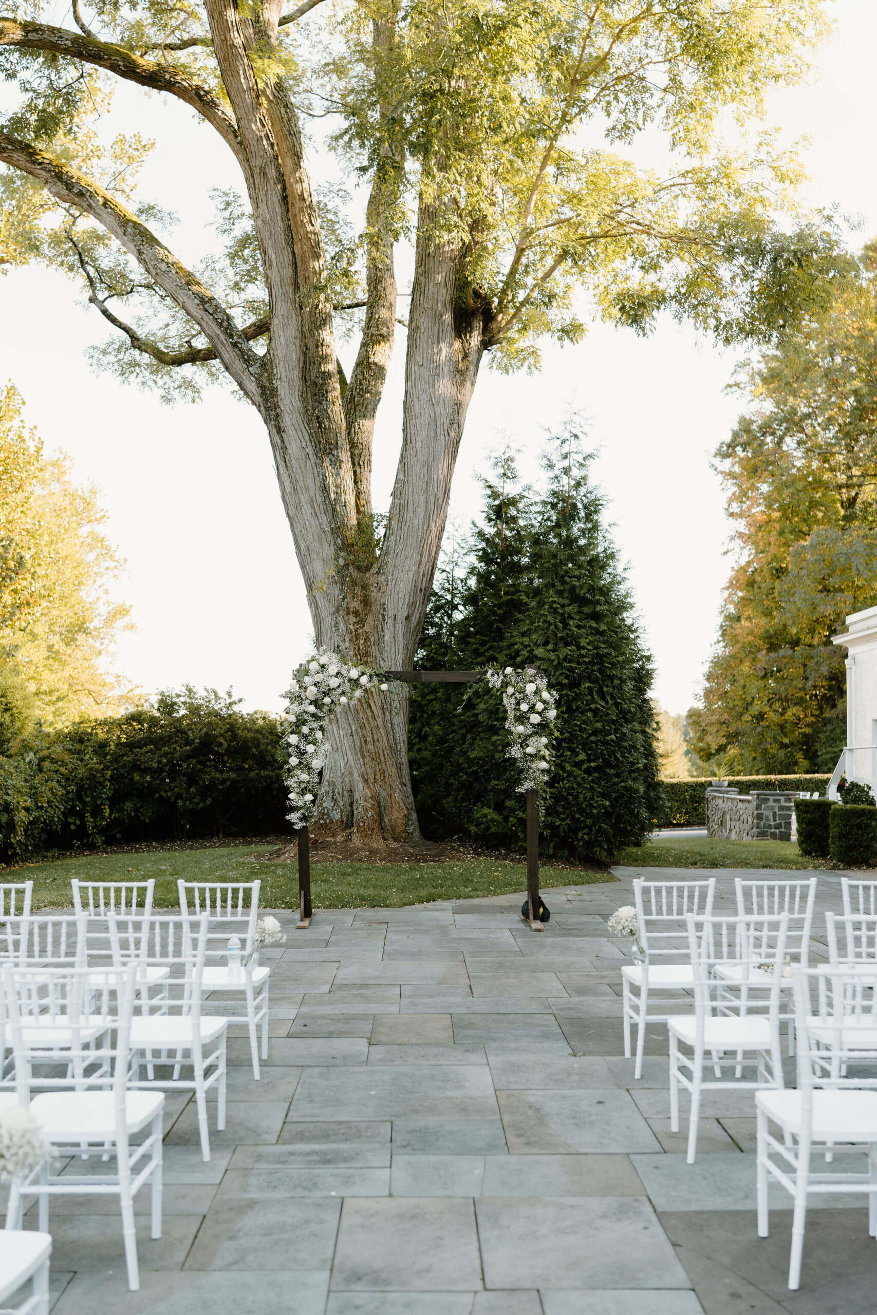 ceremony arbor underneath a large tree, aisles of white chairs leading up to it on a stone patio