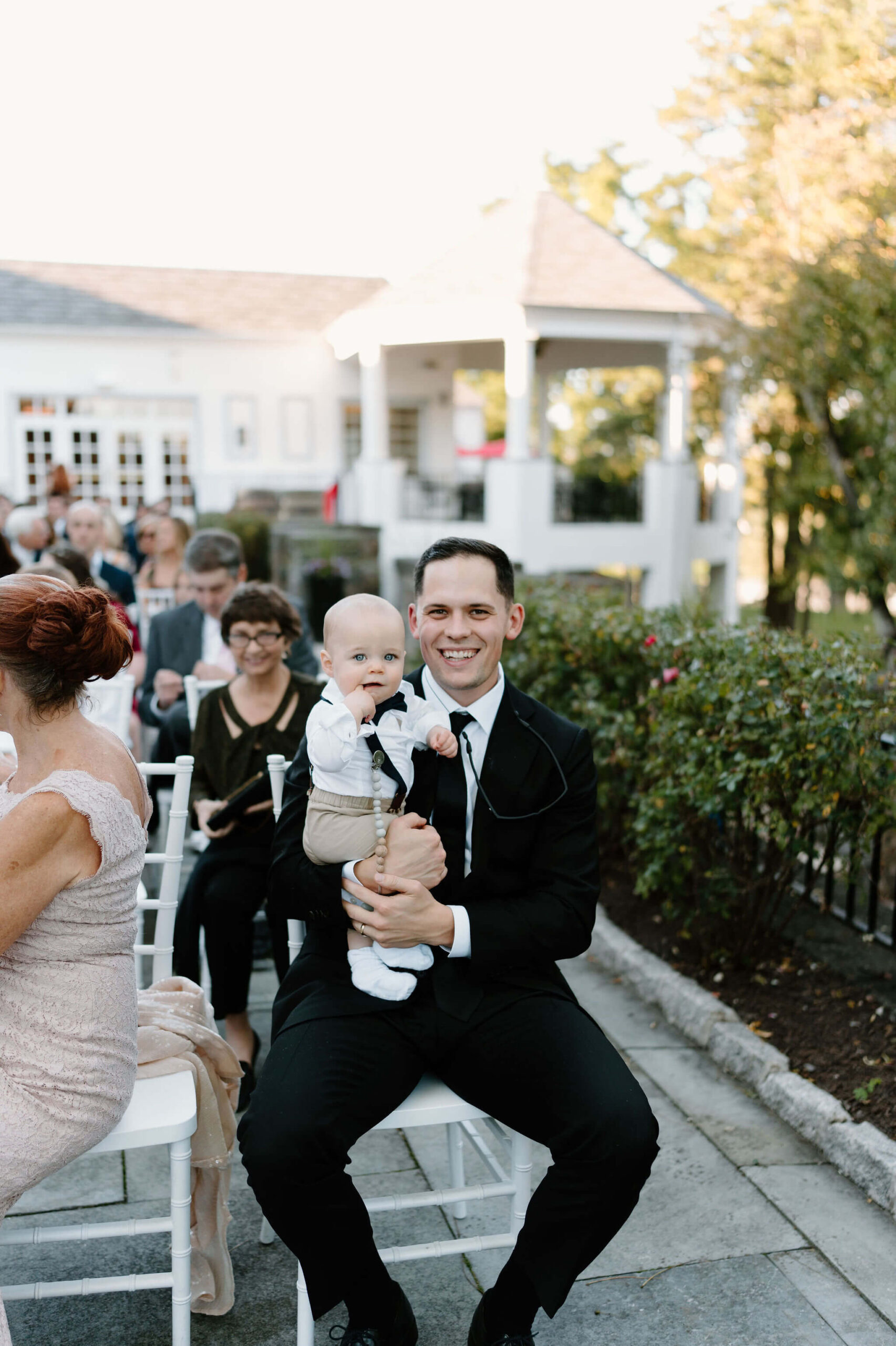 guest in a black suit holding his baby (gray khakis, white button down), both smiling at the camera