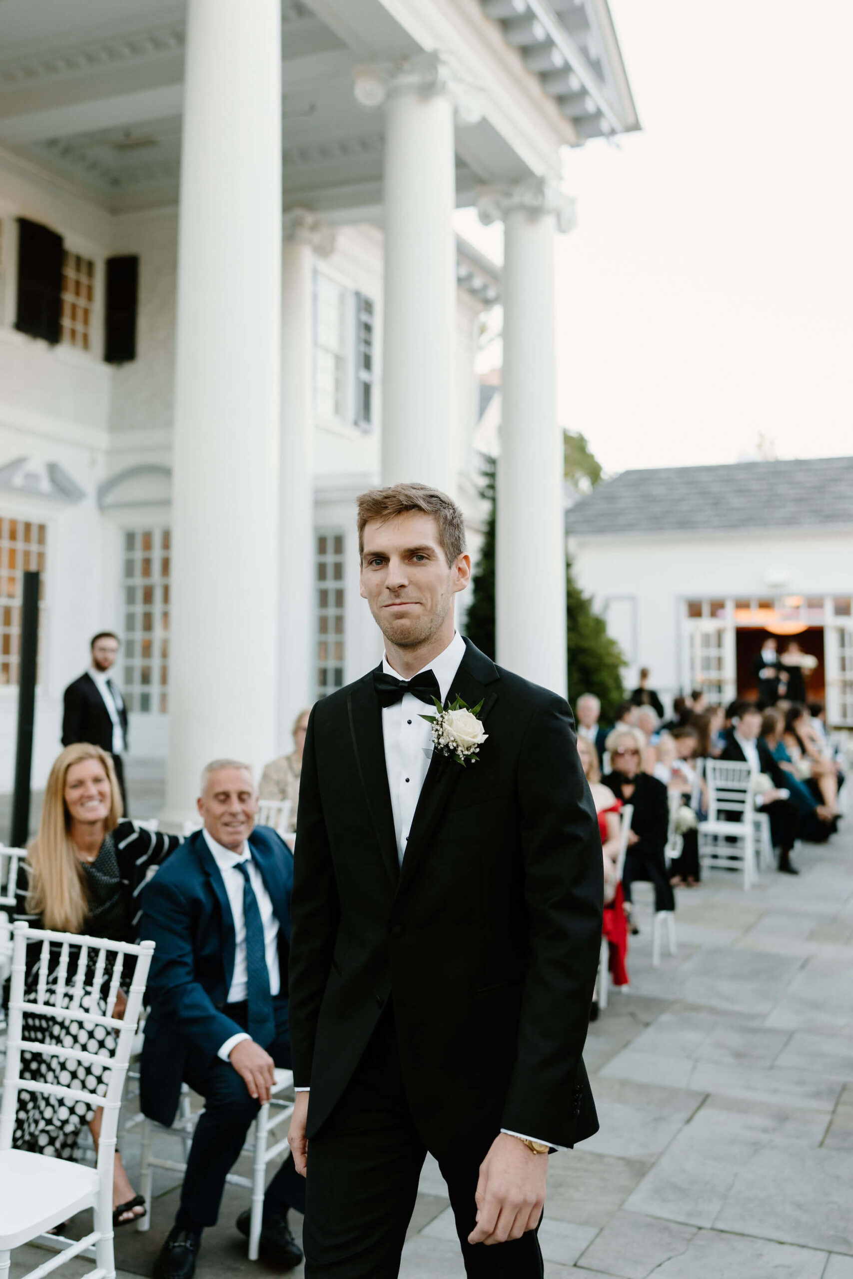 Groom smiling softly at the camera while walking down the aisle to the ceremony, guests smiling behind him