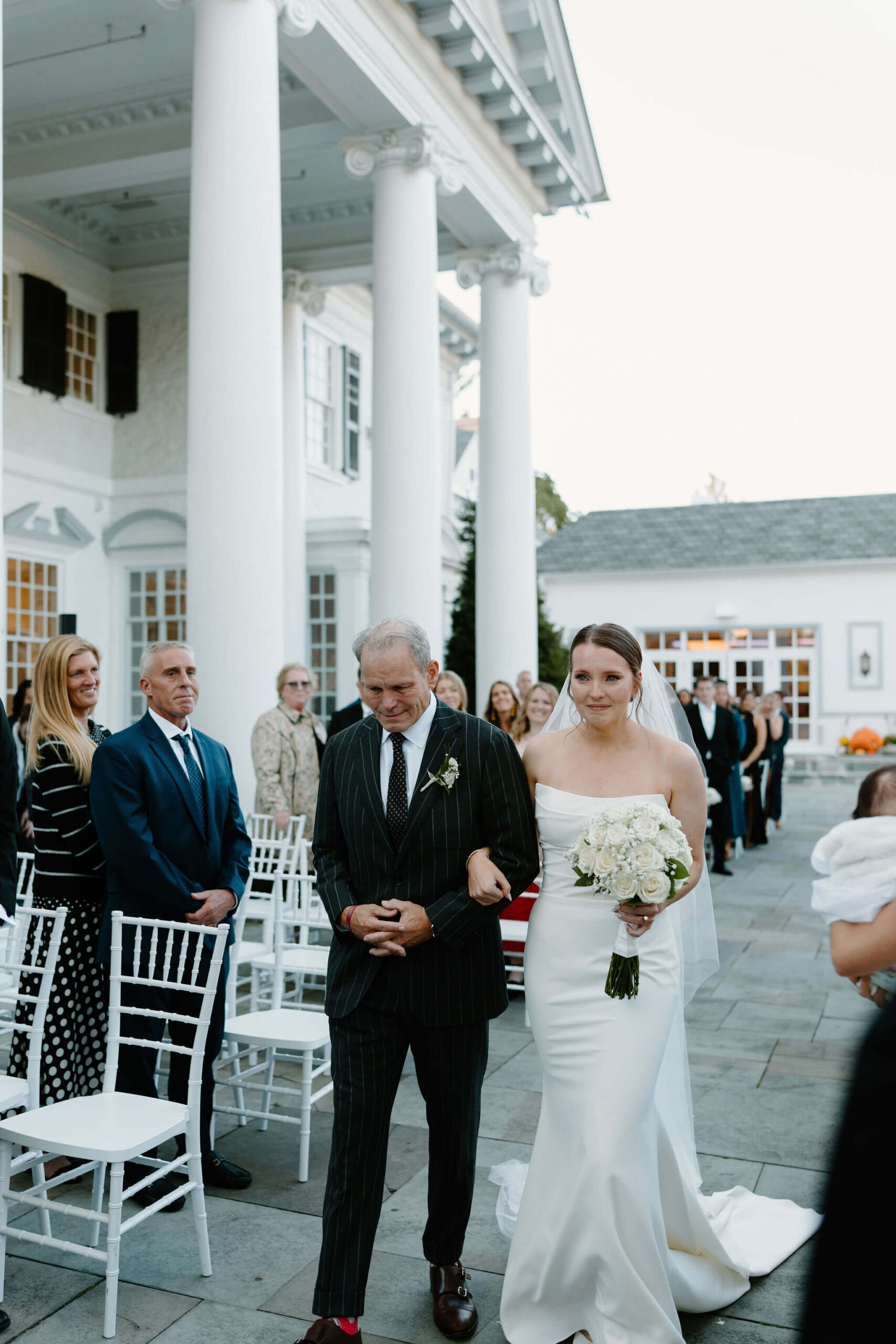 Bride and her father walking down the aisle to her wedding ceremony, both emotional while guests stand smiling around them